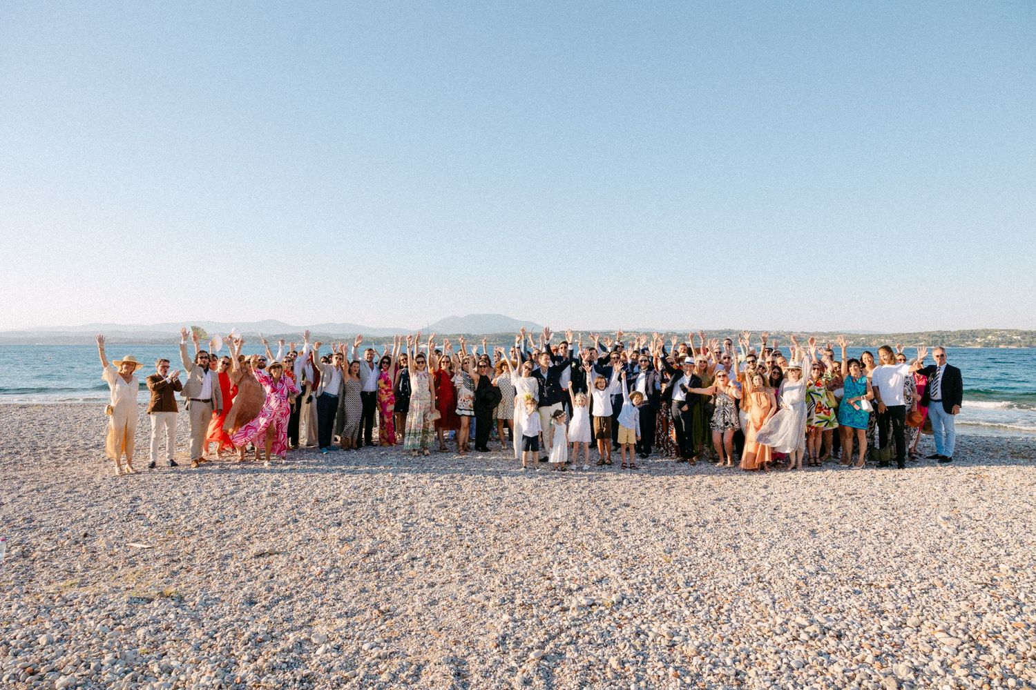 a group photo of newlyweds and their wedding guests at the beach early afternoon cheering at the camera with their hands in the air