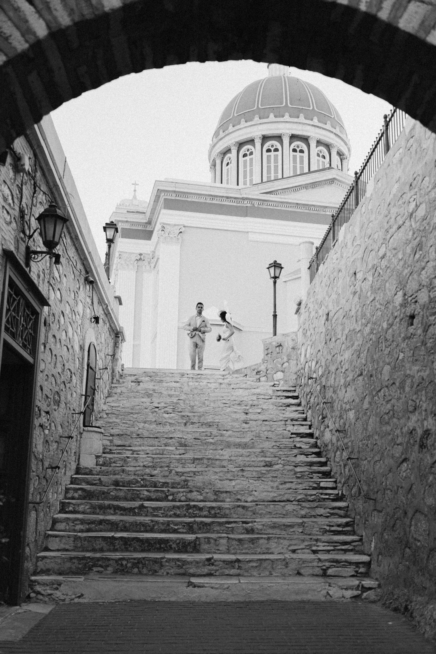 Black and white photo far away photo of a bride and groom in front of a church in Syros, Greece with wind blowing