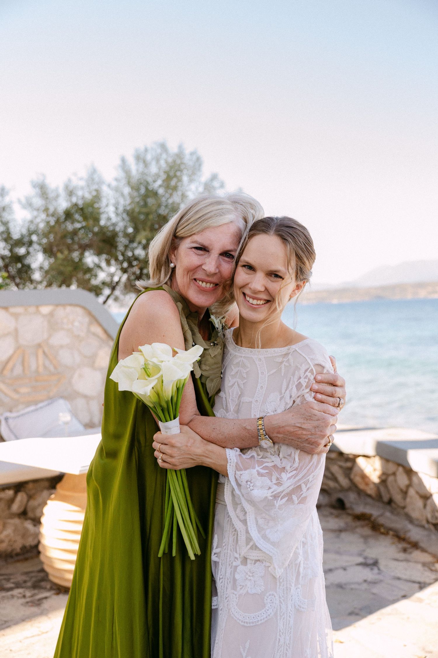 a woman wearing a green dress and a bride hugging each other and smiling to the camera. the bride is holding a bouquet of white lillies