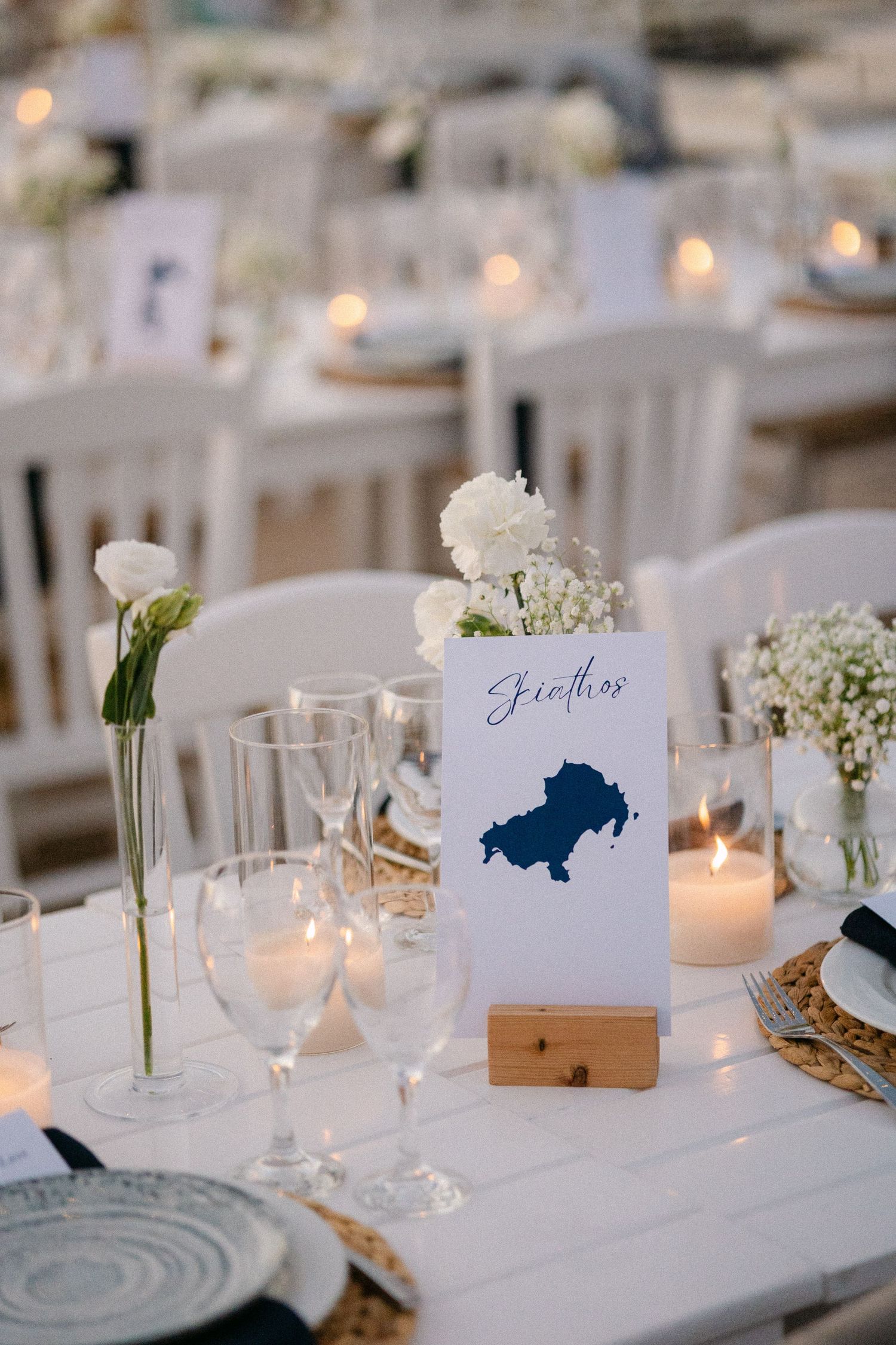 wedding table decorations showing white flowers in a vase, lit candles, blue plates and glass wines and a table sign that says: Skiathos