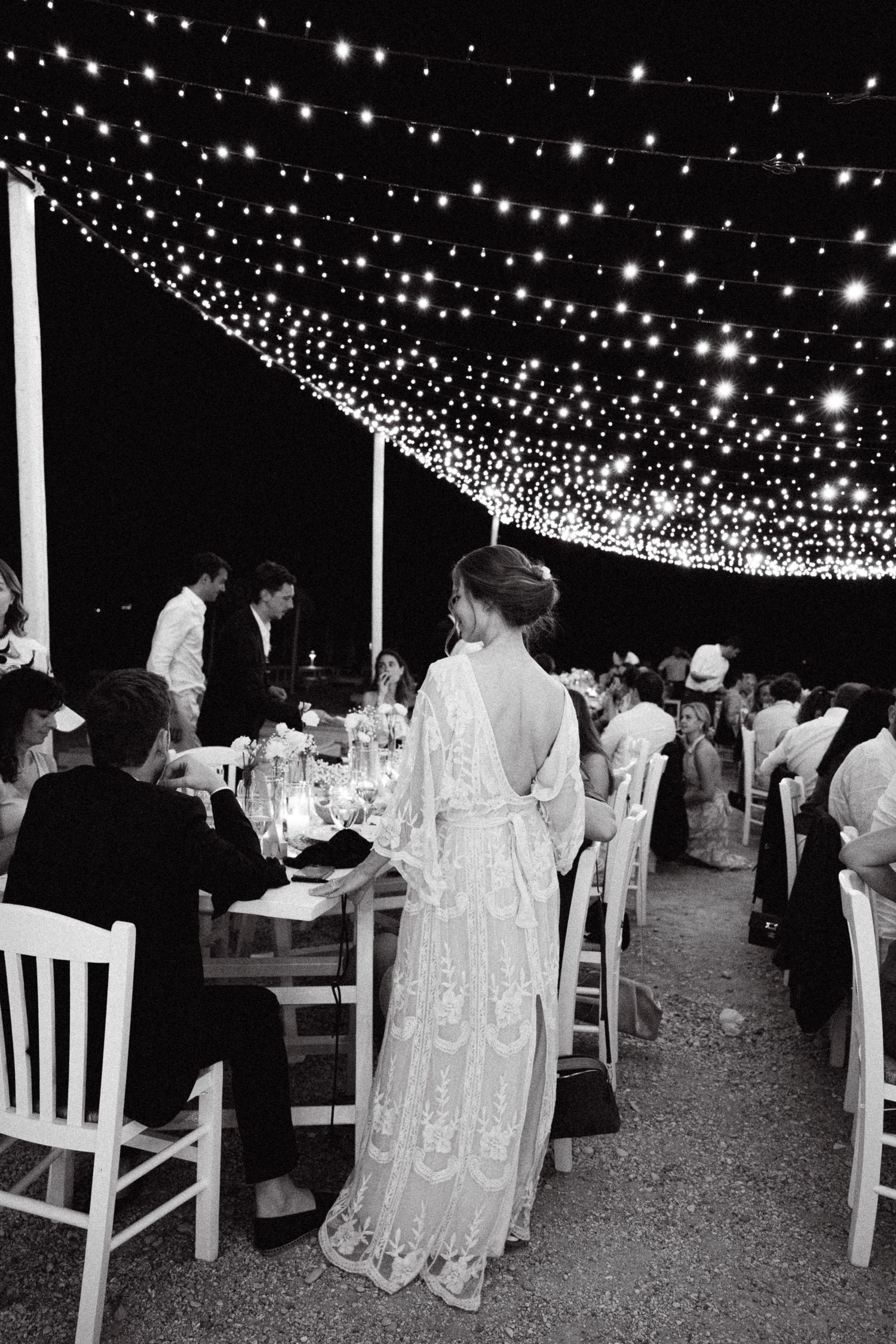 a black and white photo of a bride standing with her back on the camera chatting with wedding guests at her wedding reception in a beach bar