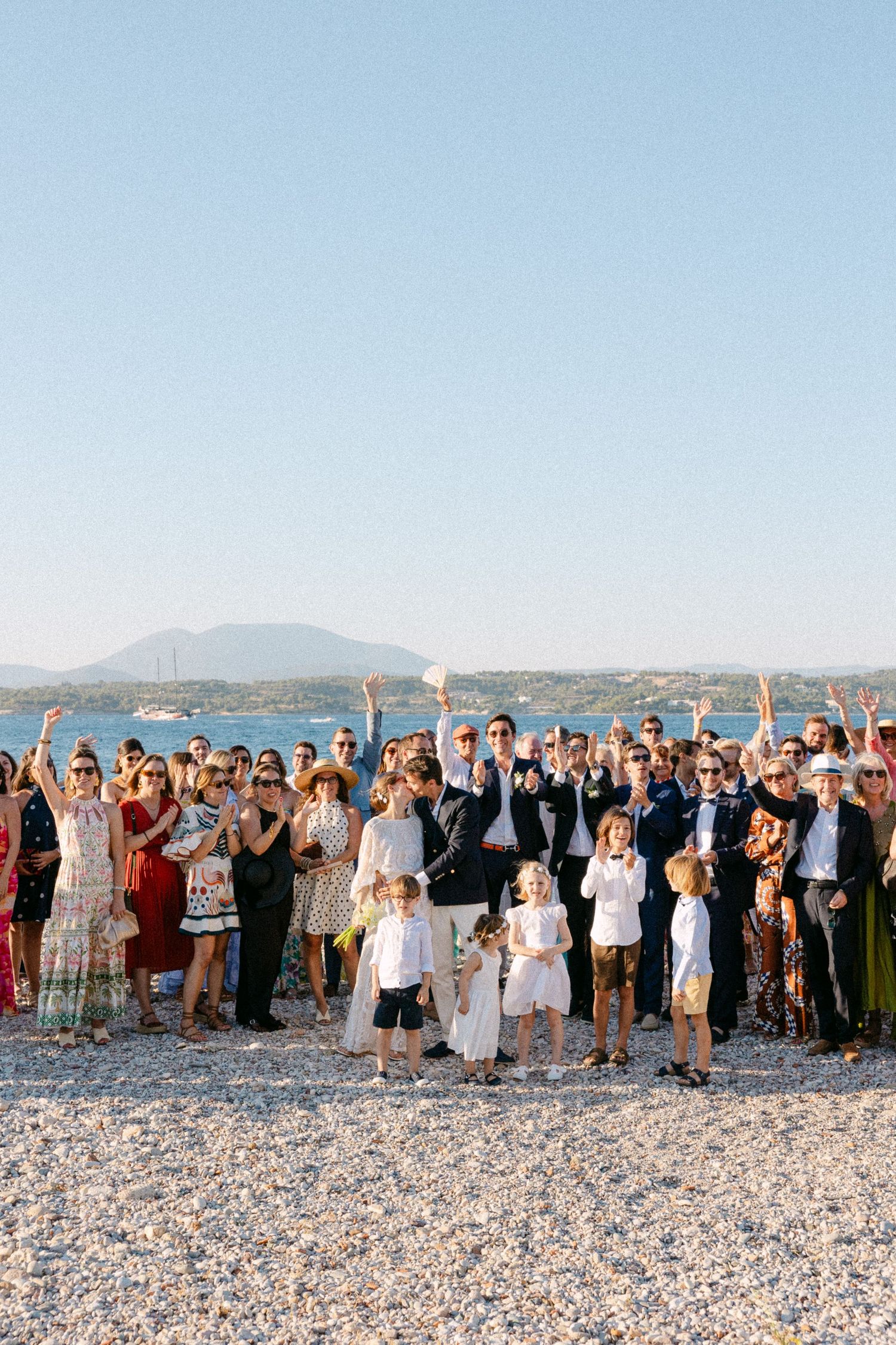 a group photo of newlyweds and their wedding guests at the beach early afternoon cheering at the camera with their hands in the air