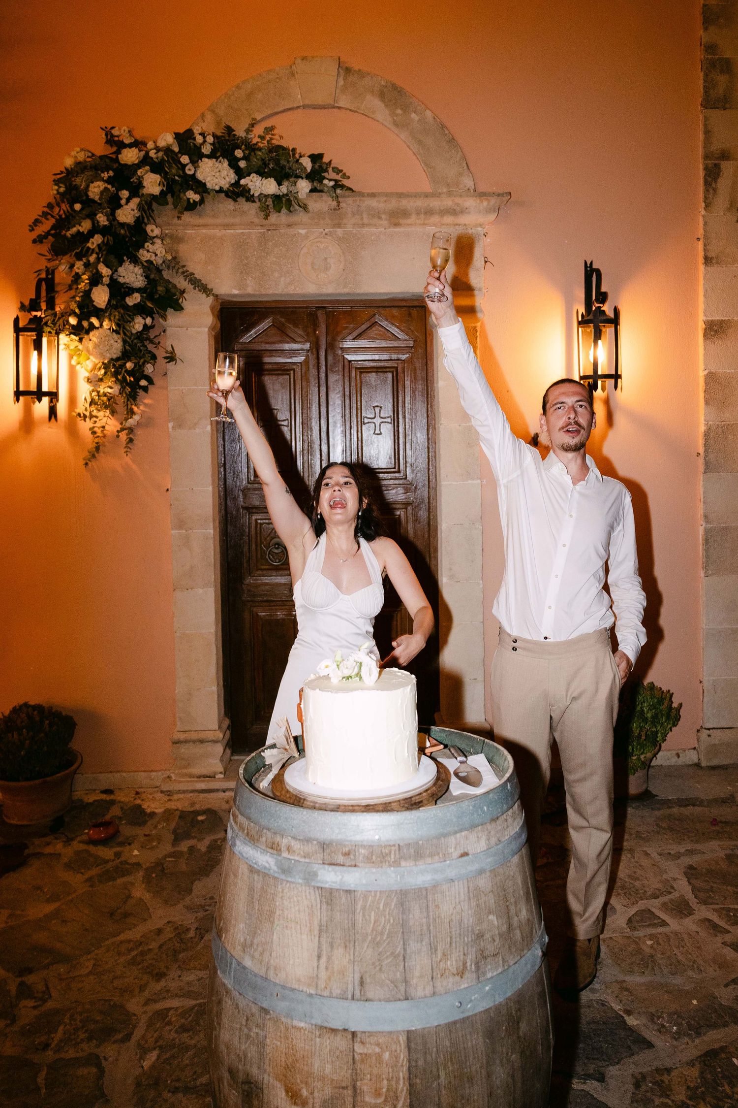 a bride and groom toasting before cutting their wedding cake at night in front of a chapel decorated with white flowers and greenery