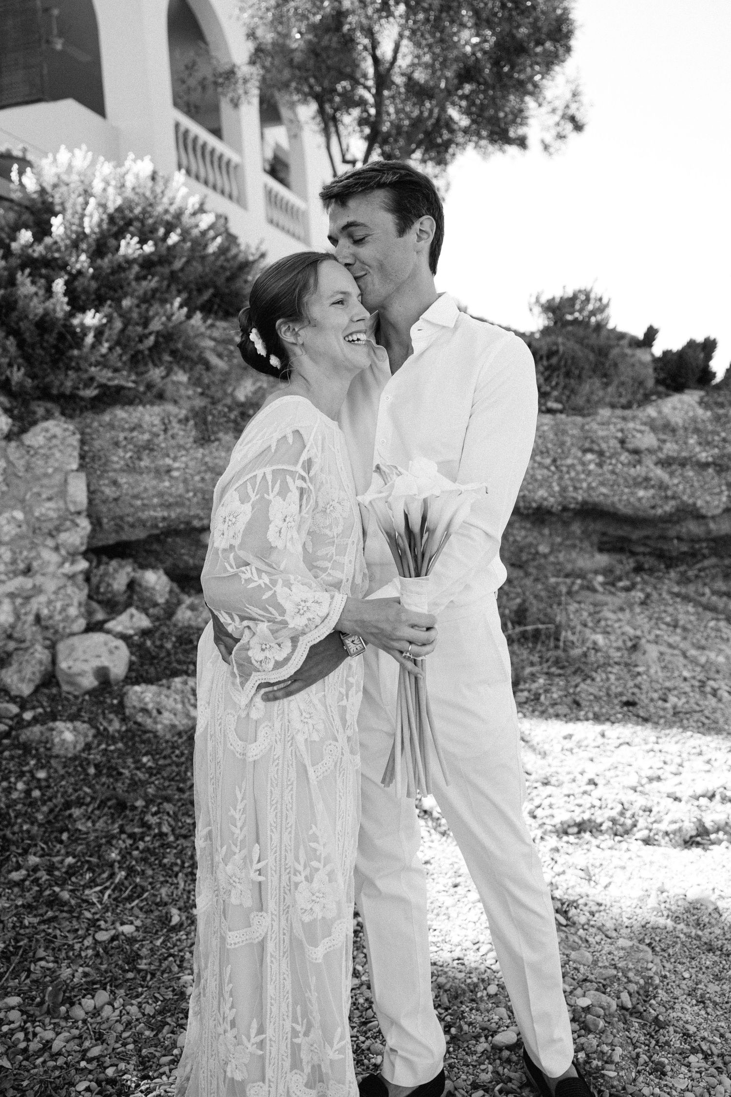 a black and white photo of a groom holding his bride and kissing her cheek in a beach setting