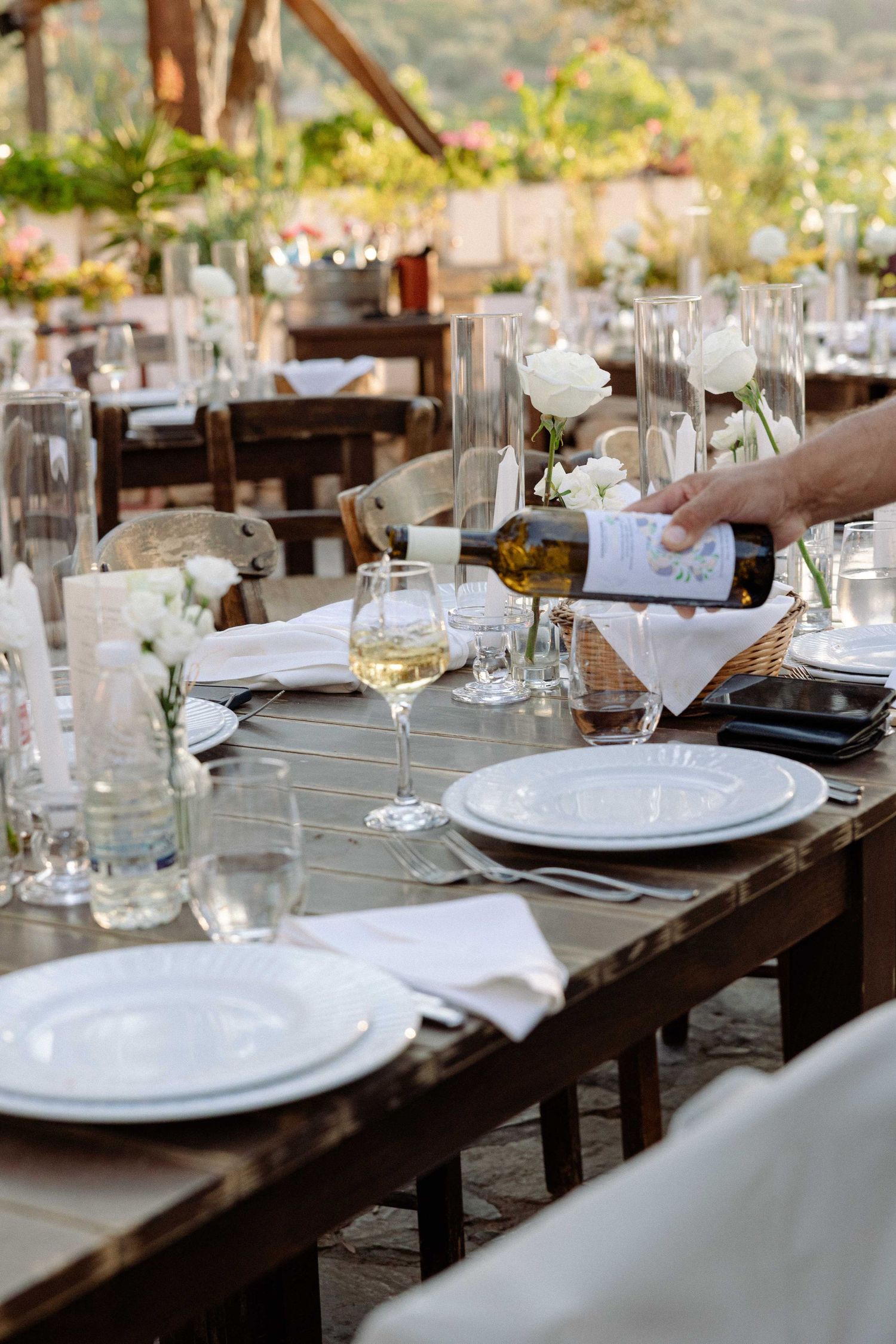 a waiter's hand as he is filling a glass with white wine in a table of wedding decorations of white roses, white plates on a wooden table