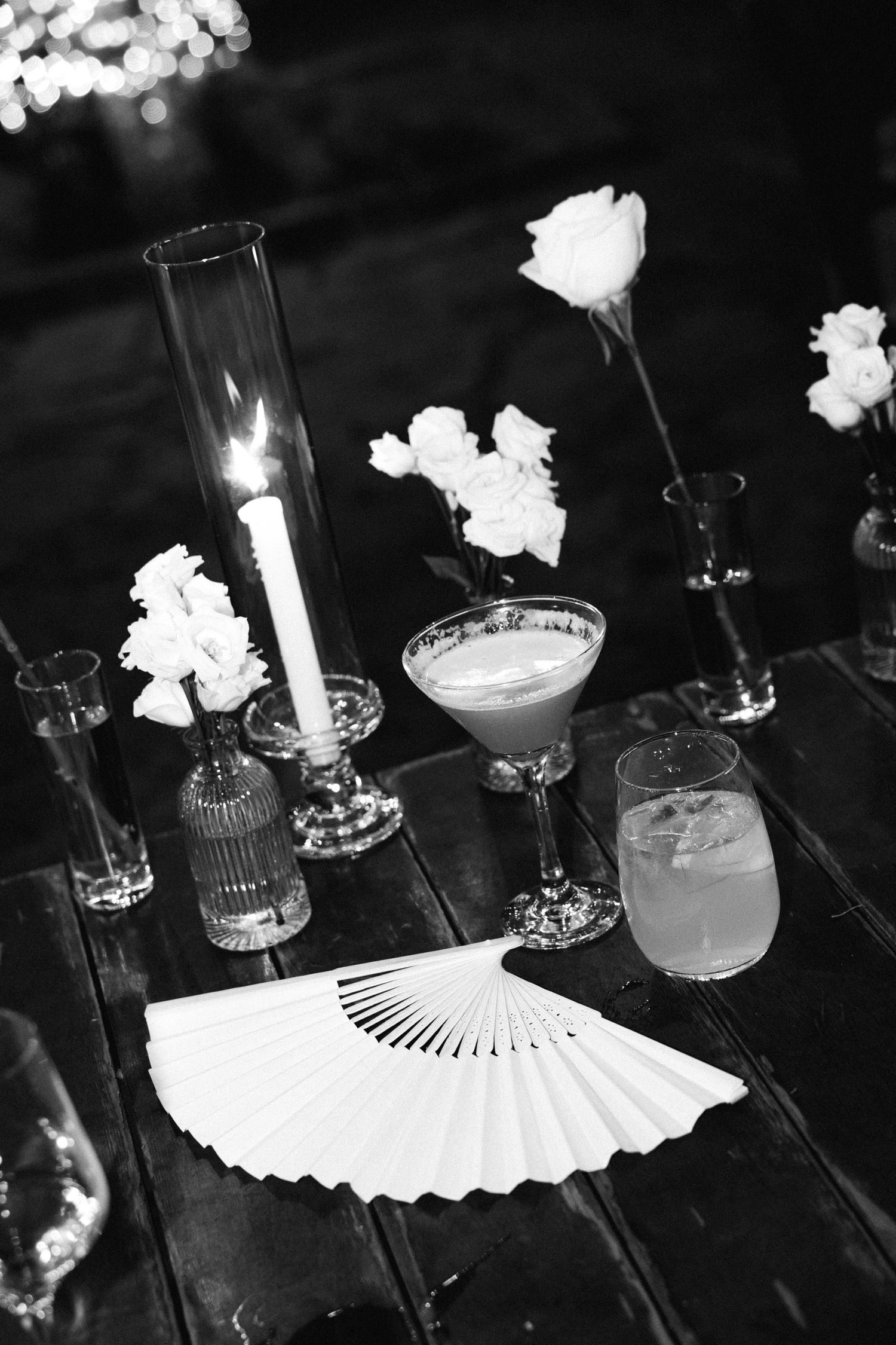 a black and white photo showing wedding details such as a white fan, vases with one white rose each, some drinks, on a wooden table