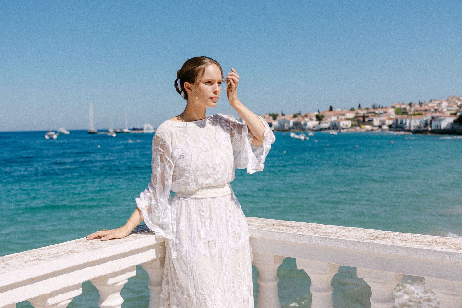a bride with an up hair do and a lace long-sleeve wedding dress standing in a balcony with the sea as a background in a sunny setting