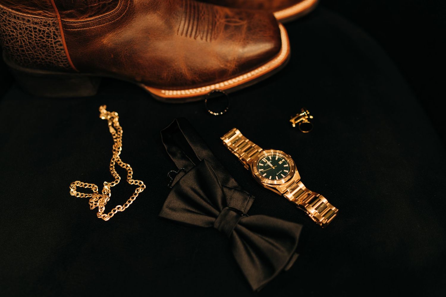 Wedding flat lay featuring gold watch, black bow tie, gold chain necklace, and cowboy boots.