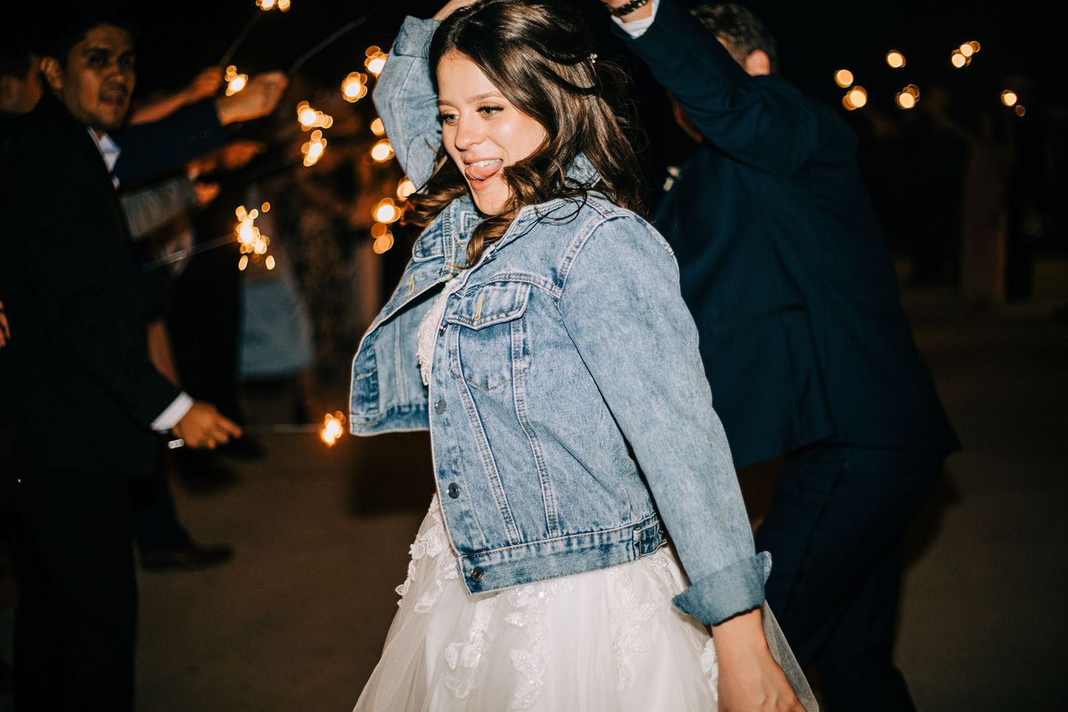 Smiling bride in a white tulle gown and denim jacket walks through a sparkler exit at night.