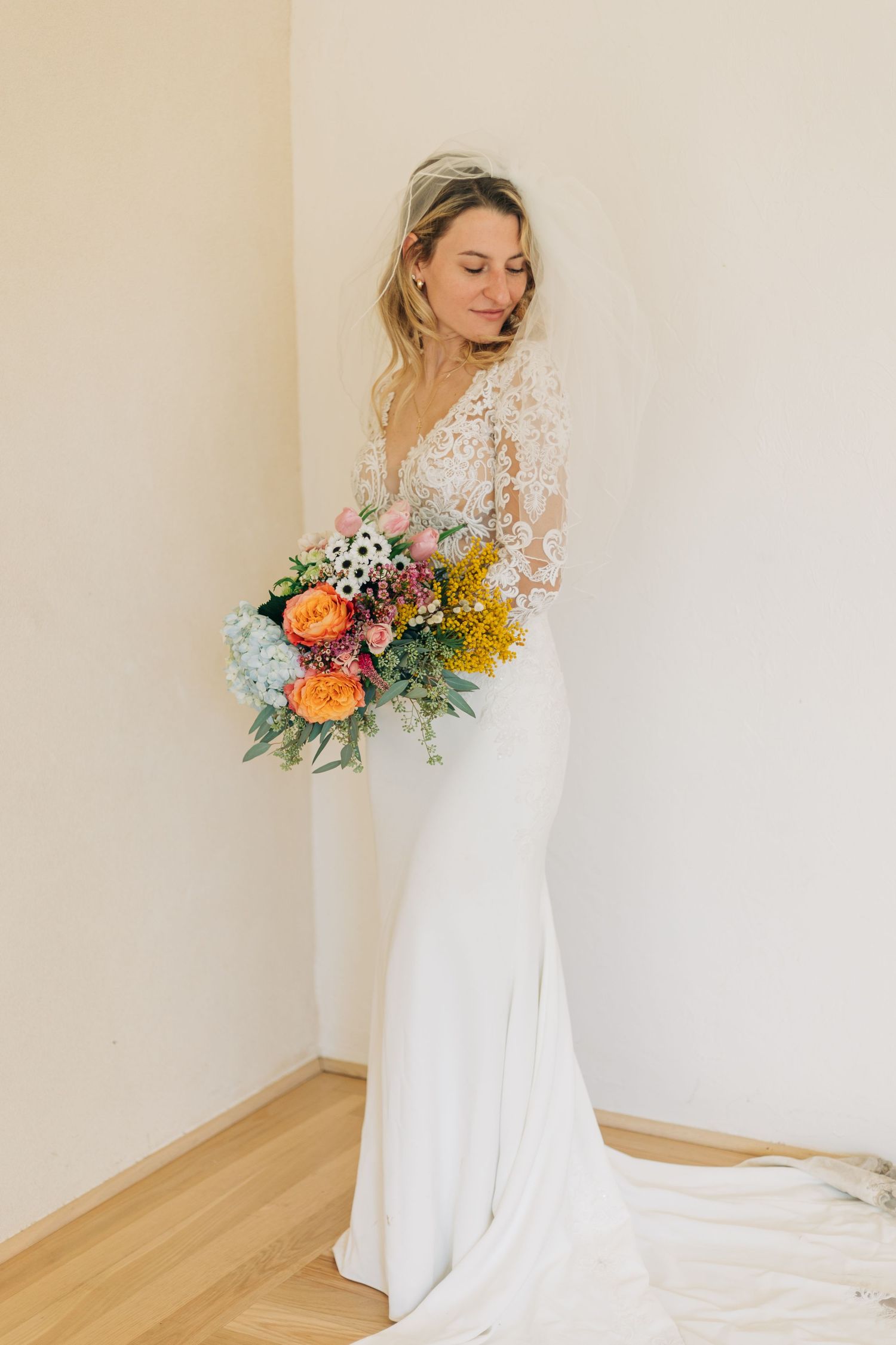 Bride in elegant white lace long-sleeve wedding gown holding a vibrant colorful bouquet indoors.