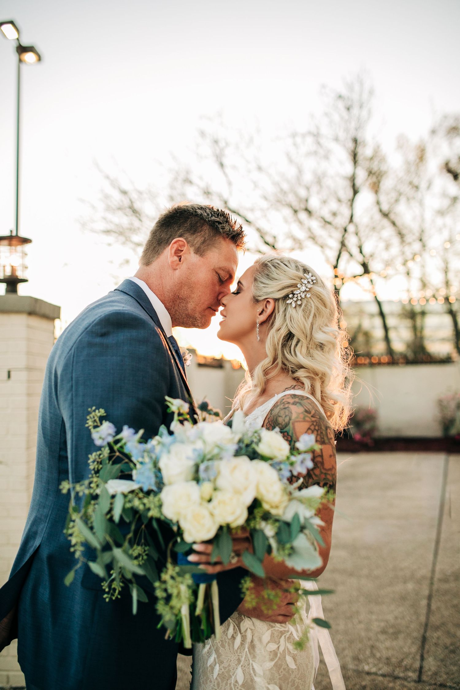 Tattooed bride and groom share a tender forehead kiss at sunset, bride holding a lush white floral bouquet.