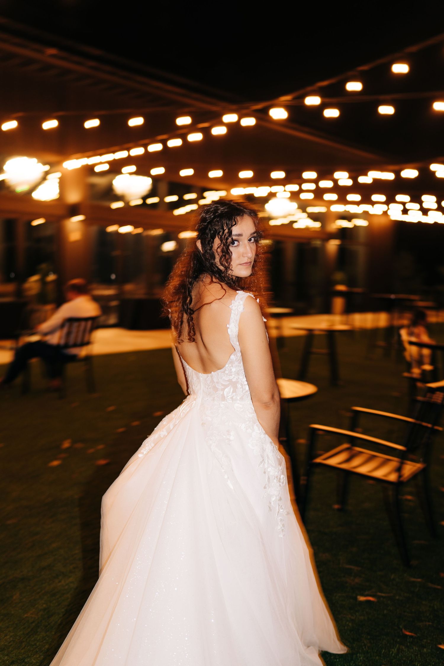 Elegant bride in a backless lace gown posing under warm string lights at an outdoor evening reception.