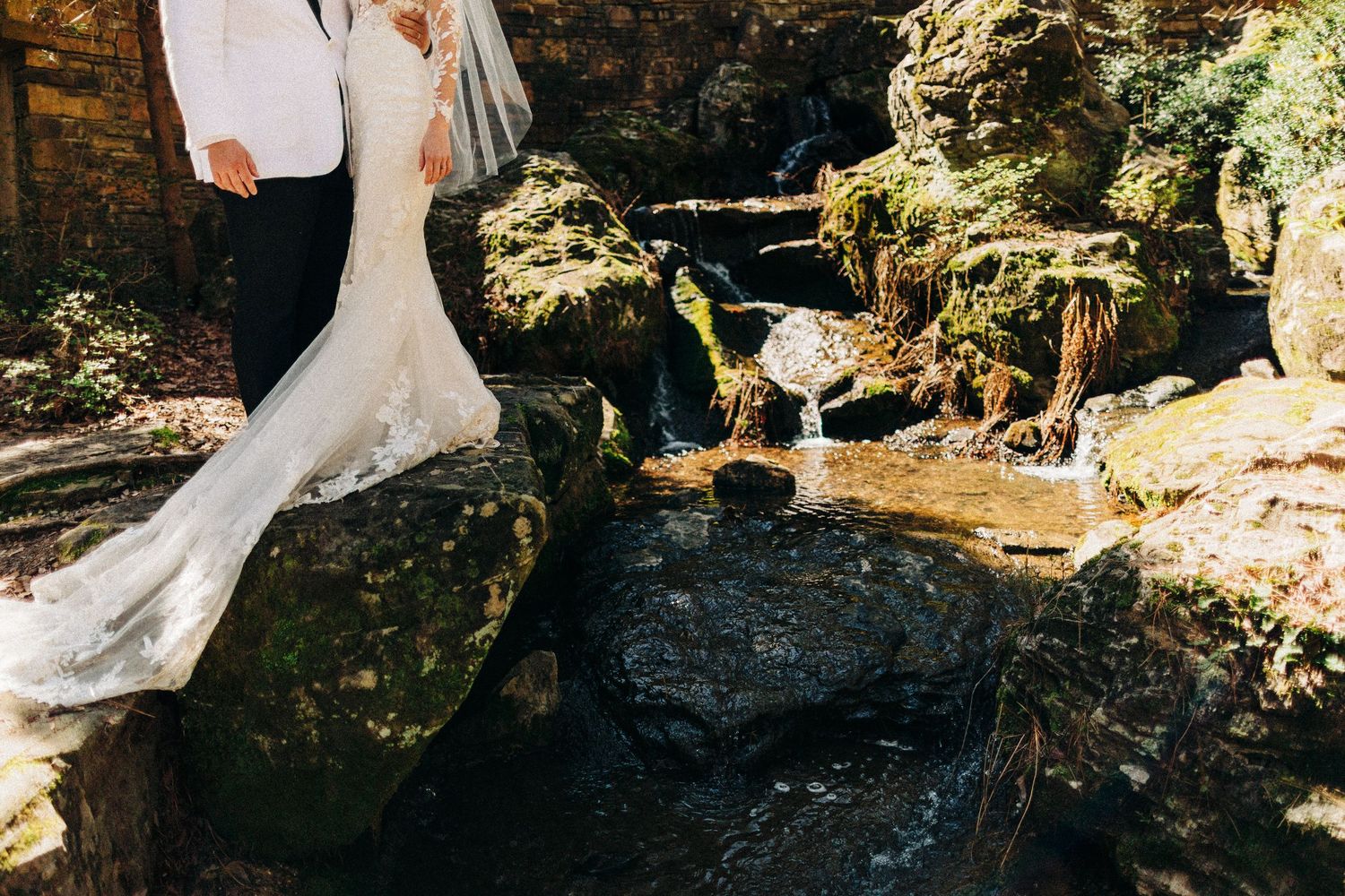 Wedding couple standing on mossy rocks beside a serene waterfall in a lush outdoor setting.