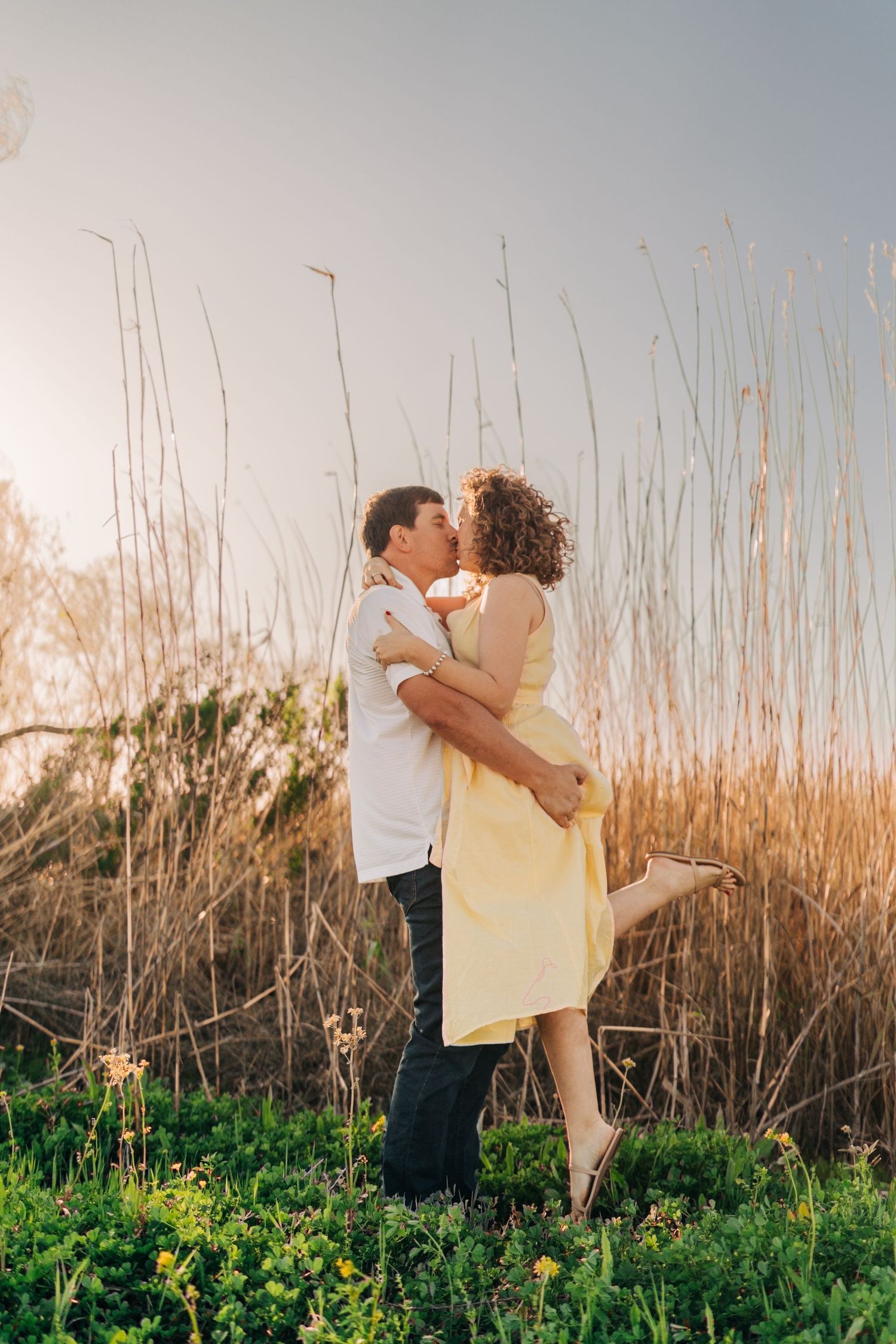 Couple sharing a romantic kiss outdoors at sunset, man lifting woman in yellow dress among tall golden grasses.