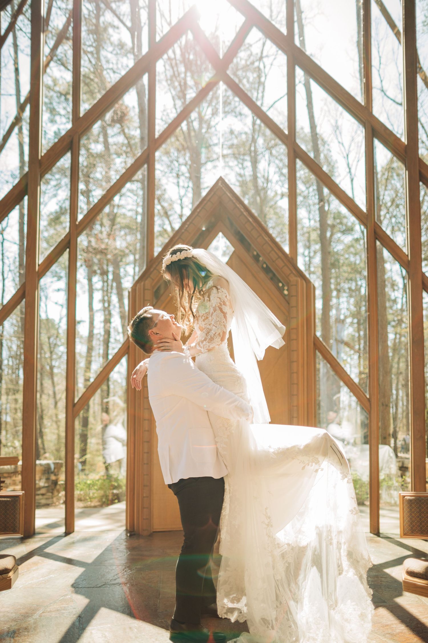 Groom lifts bride in a sunlit glass chapel surrounded by tall trees, creating a magical romantic moment.