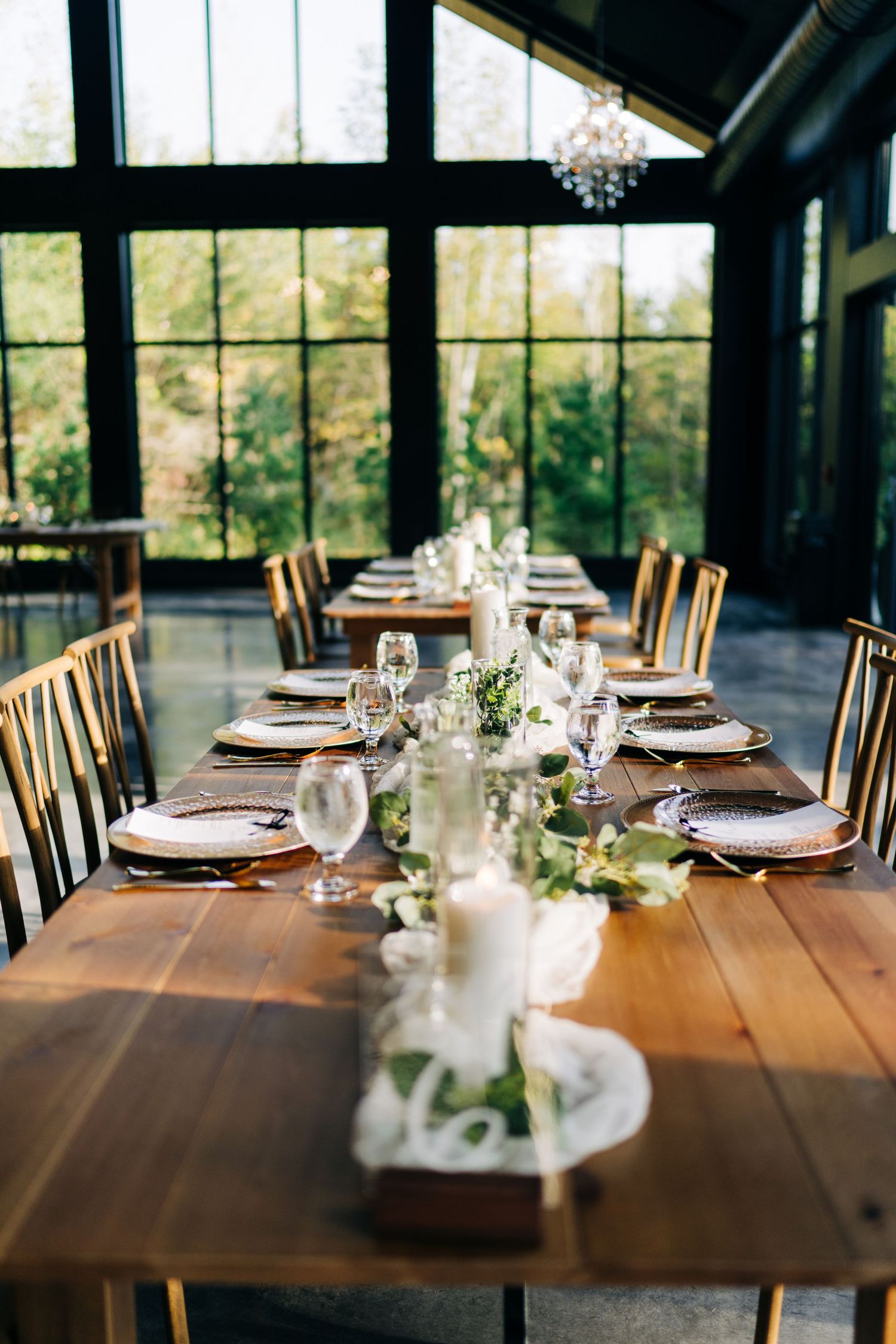 Elegant wedding reception table set with white florals, candles, crystal glasses, and gold chargers in a modern greenhouse venue.
