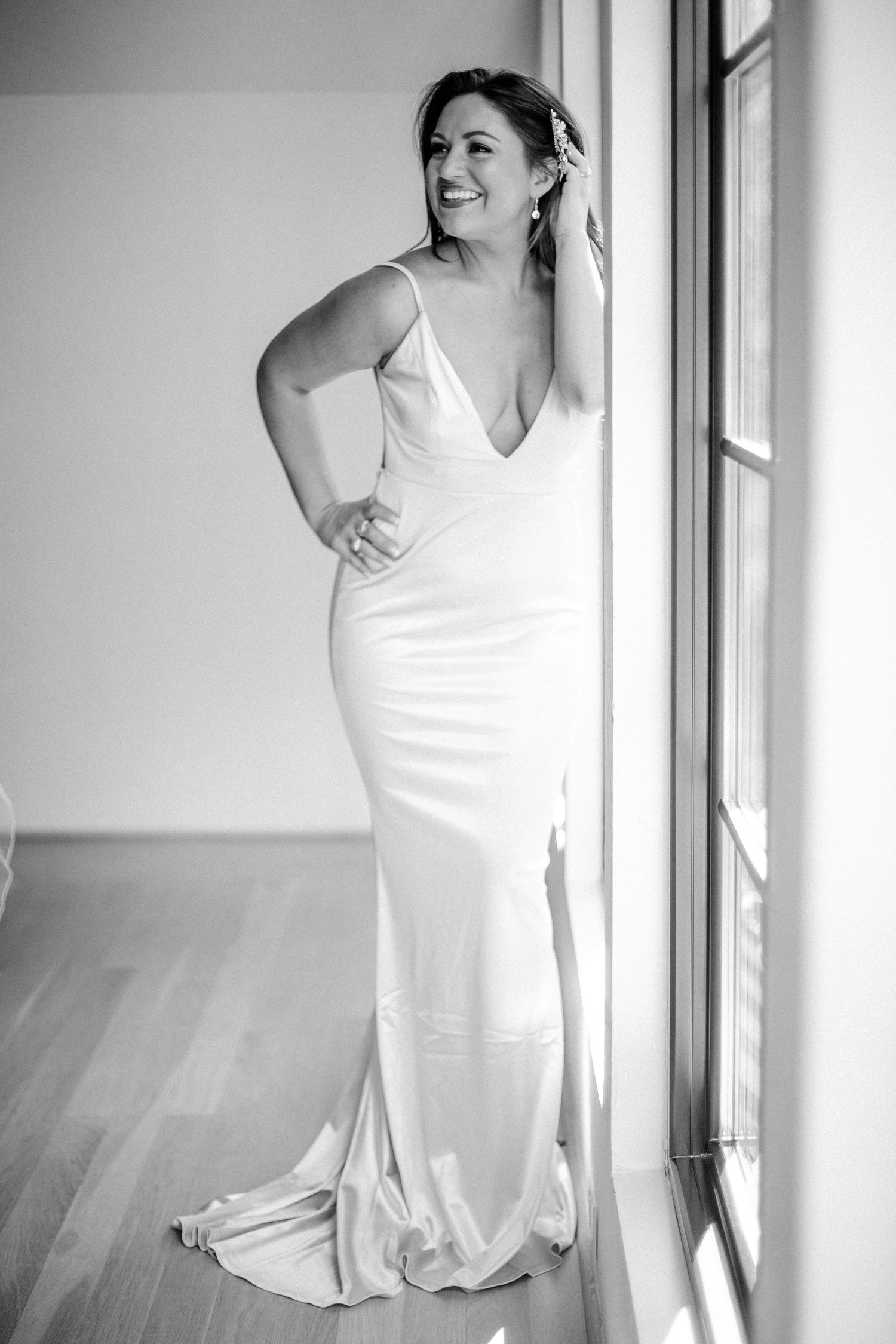 Elegant bride in sleek satin gown posing by window in black and white portrait.