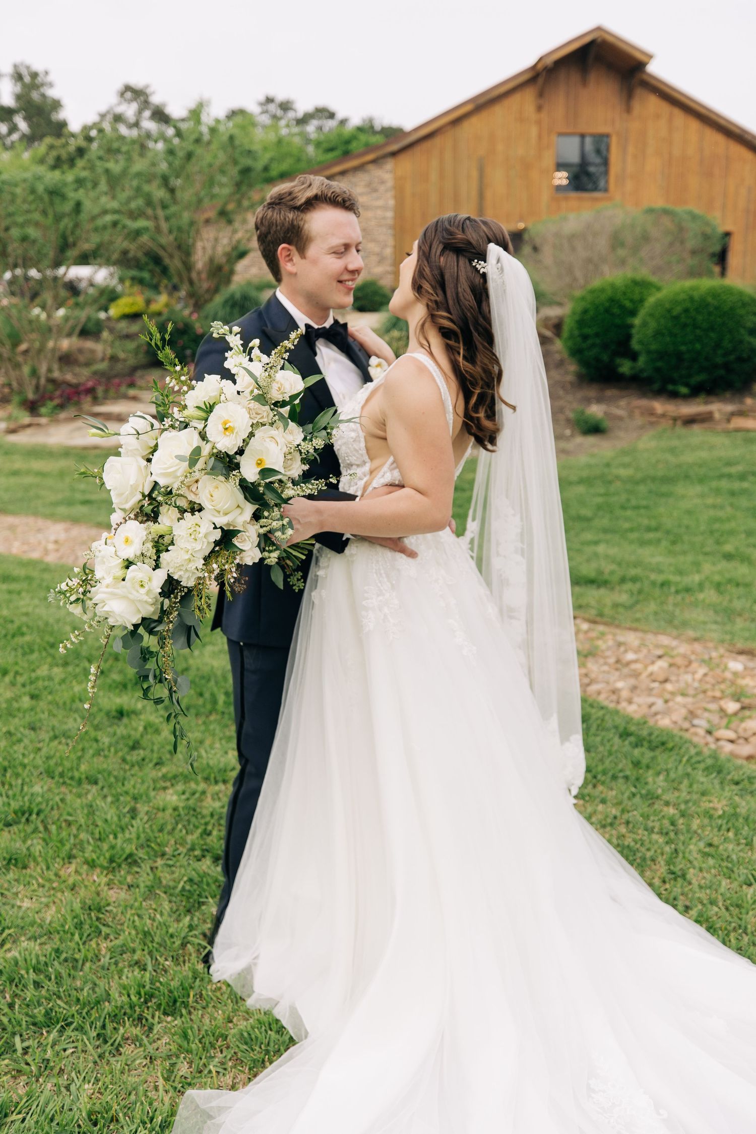 Bride and groom embracing outdoors holding white floral bouquet near rustic barn venue.