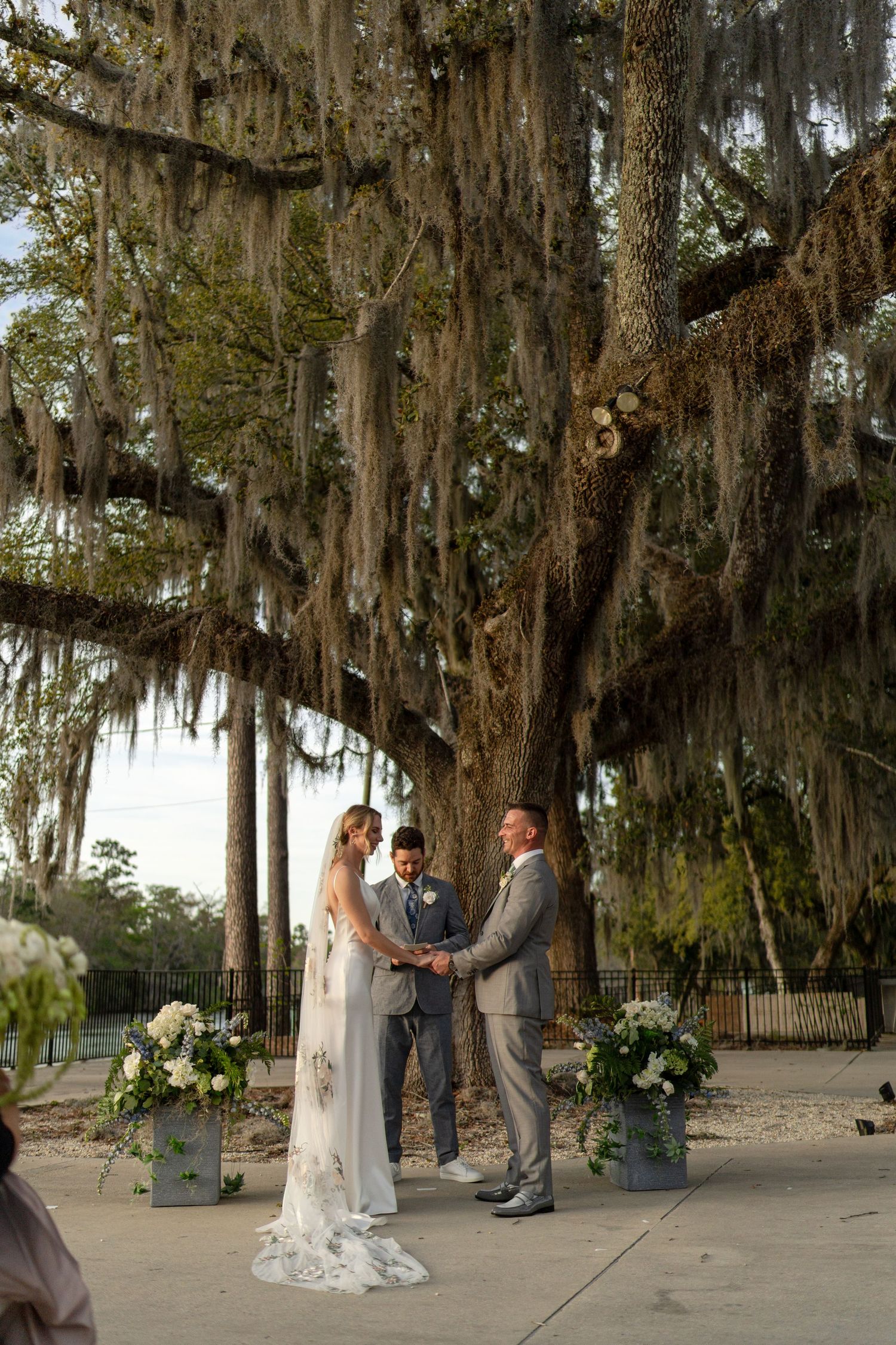 Bride and groom exchanging vows beneath massive Spanish moss-covered oak tree with white floral arrangements.