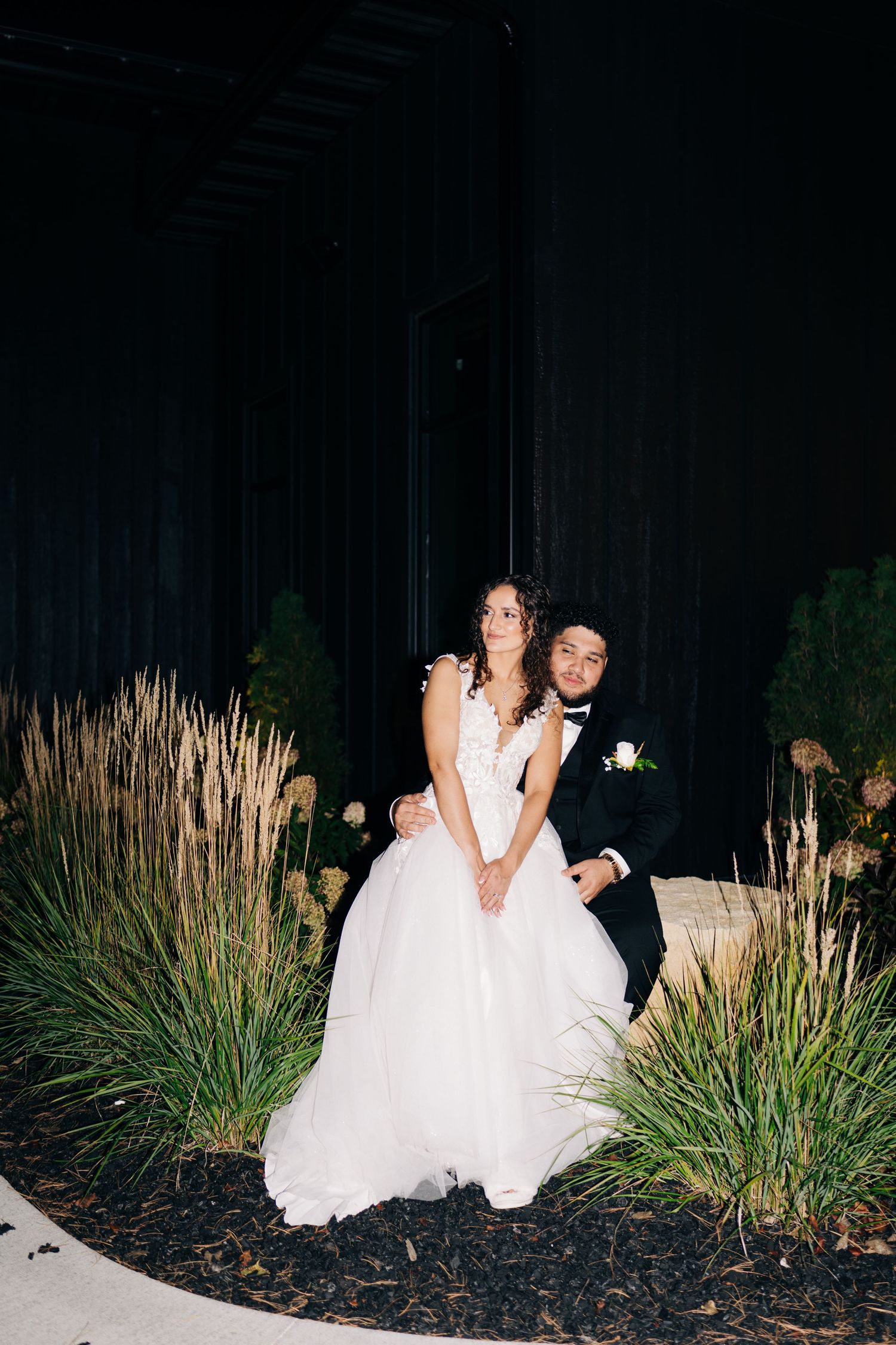 Bride and groom posing elegantly among ornamental grasses at a dark, dramatic indoor wedding venue.