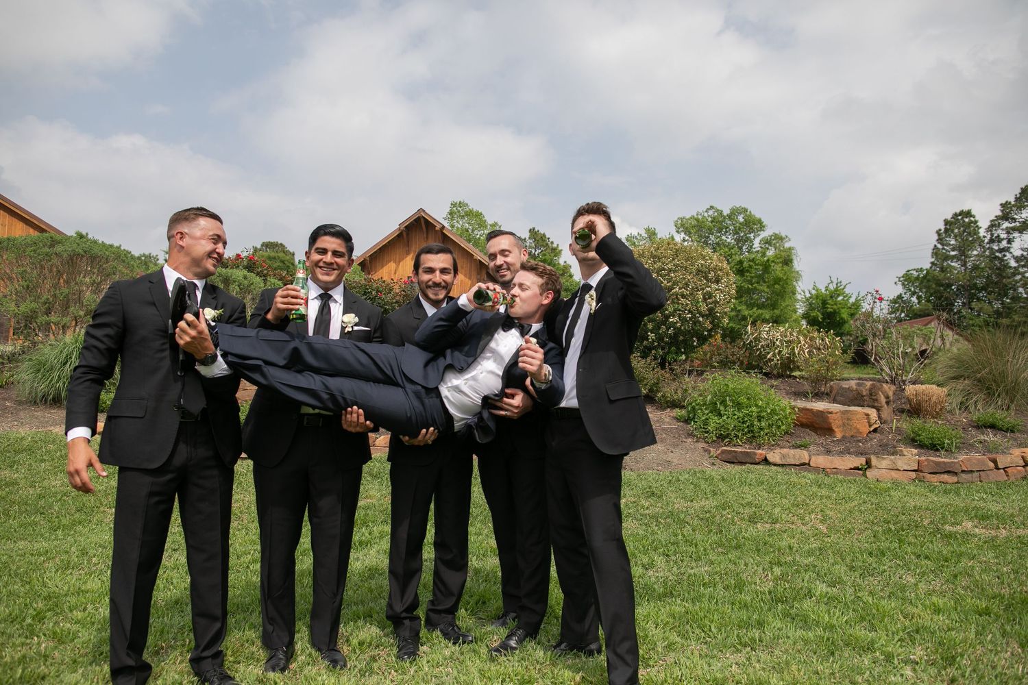 Groomsmen playfully lift the groom outdoors on a grassy lawn in matching black suits before the wedding.