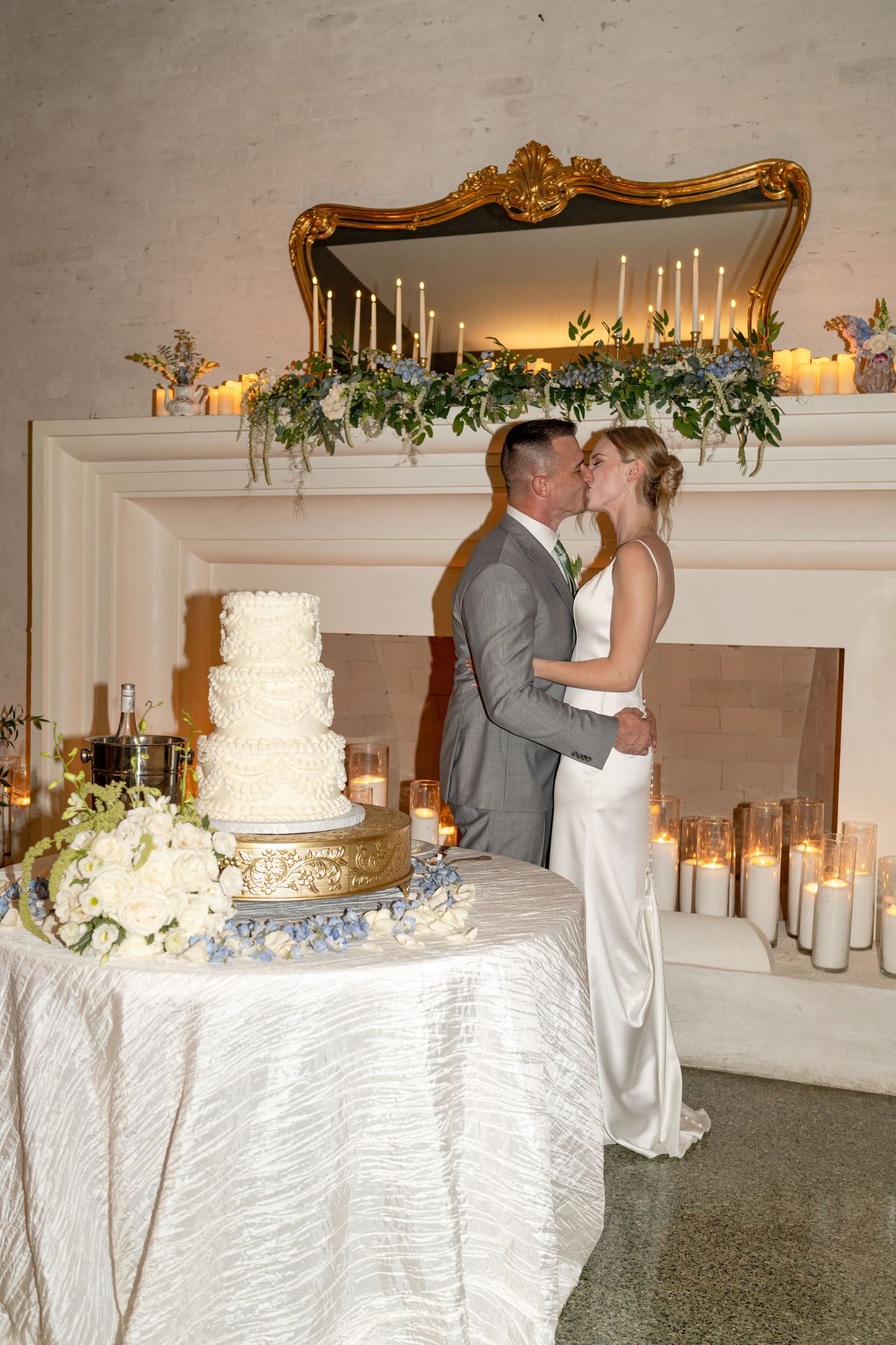 Bride and groom kiss beside a three-tier wedding cake near a candlelit fireplace mantle.