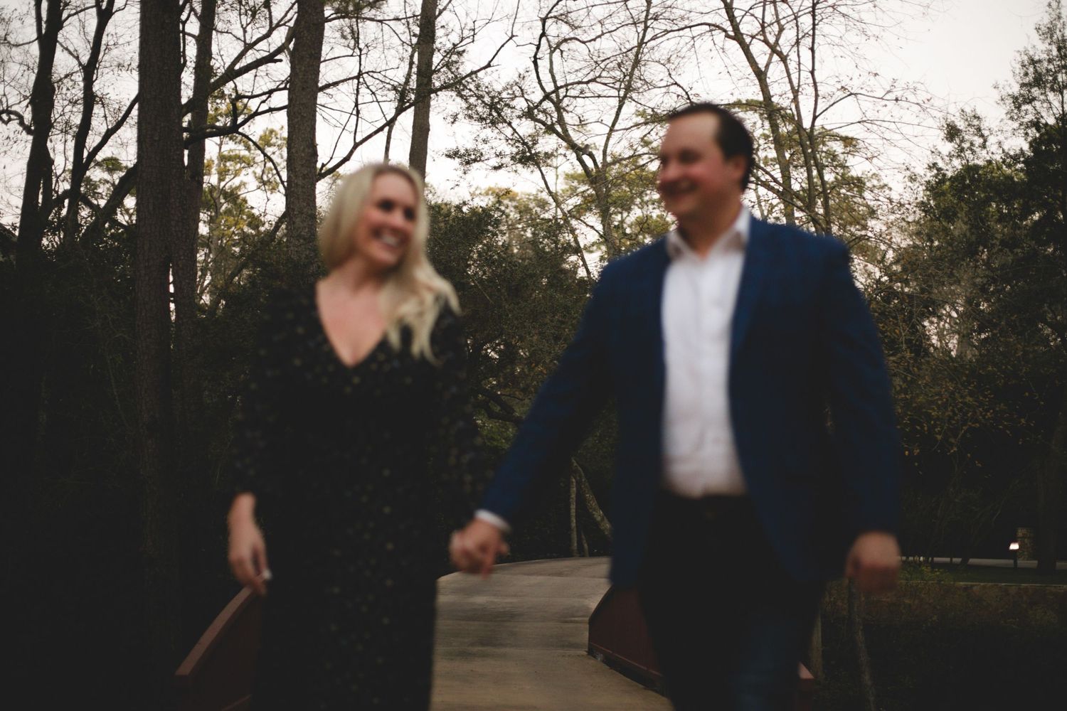 Couple walks hand-in-hand along wooded path during romantic outdoor engagement session at dusk.