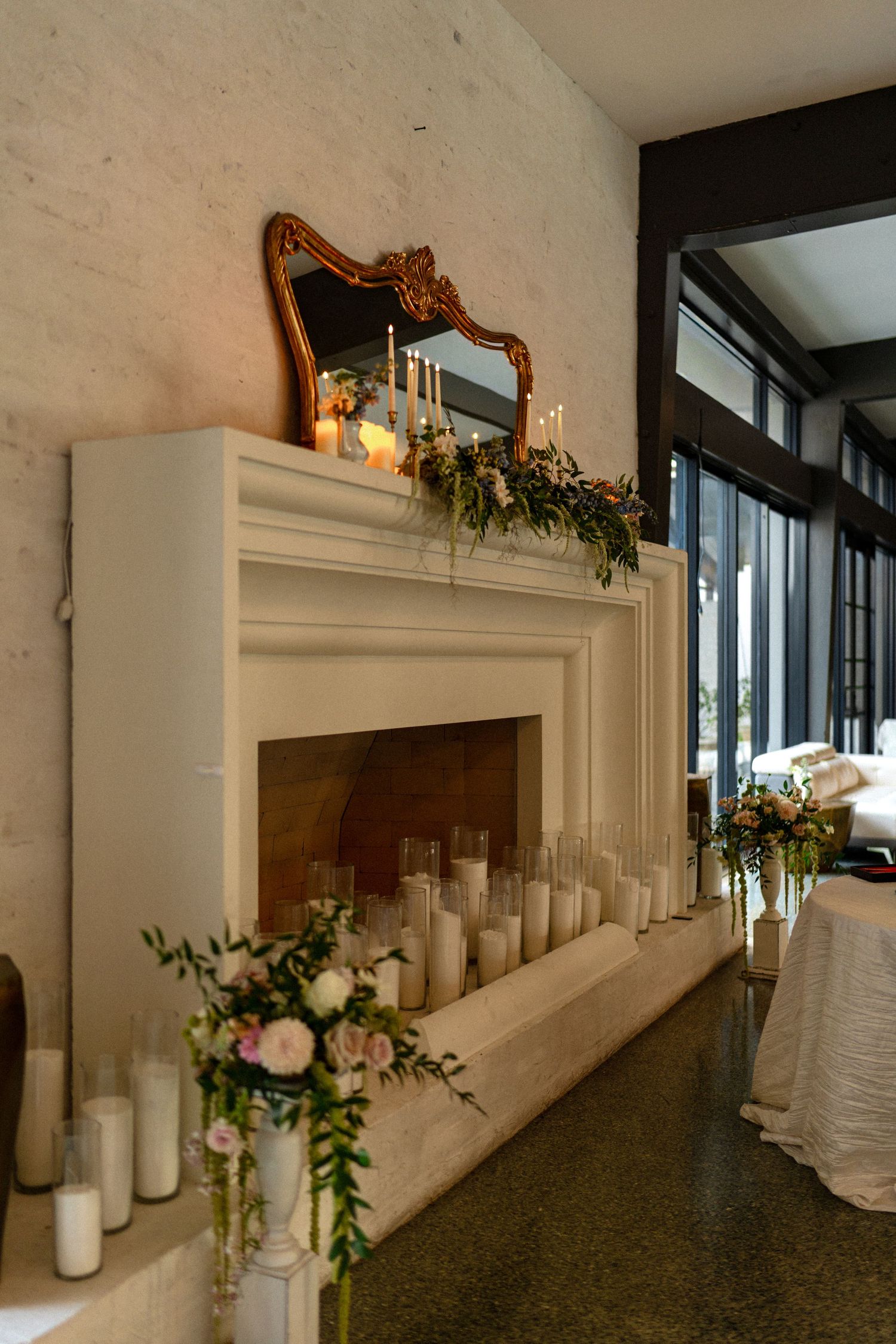 Elegant white fireplace mantel decorated with candles, flowers, and a gold ornate mirror for a wedding reception.