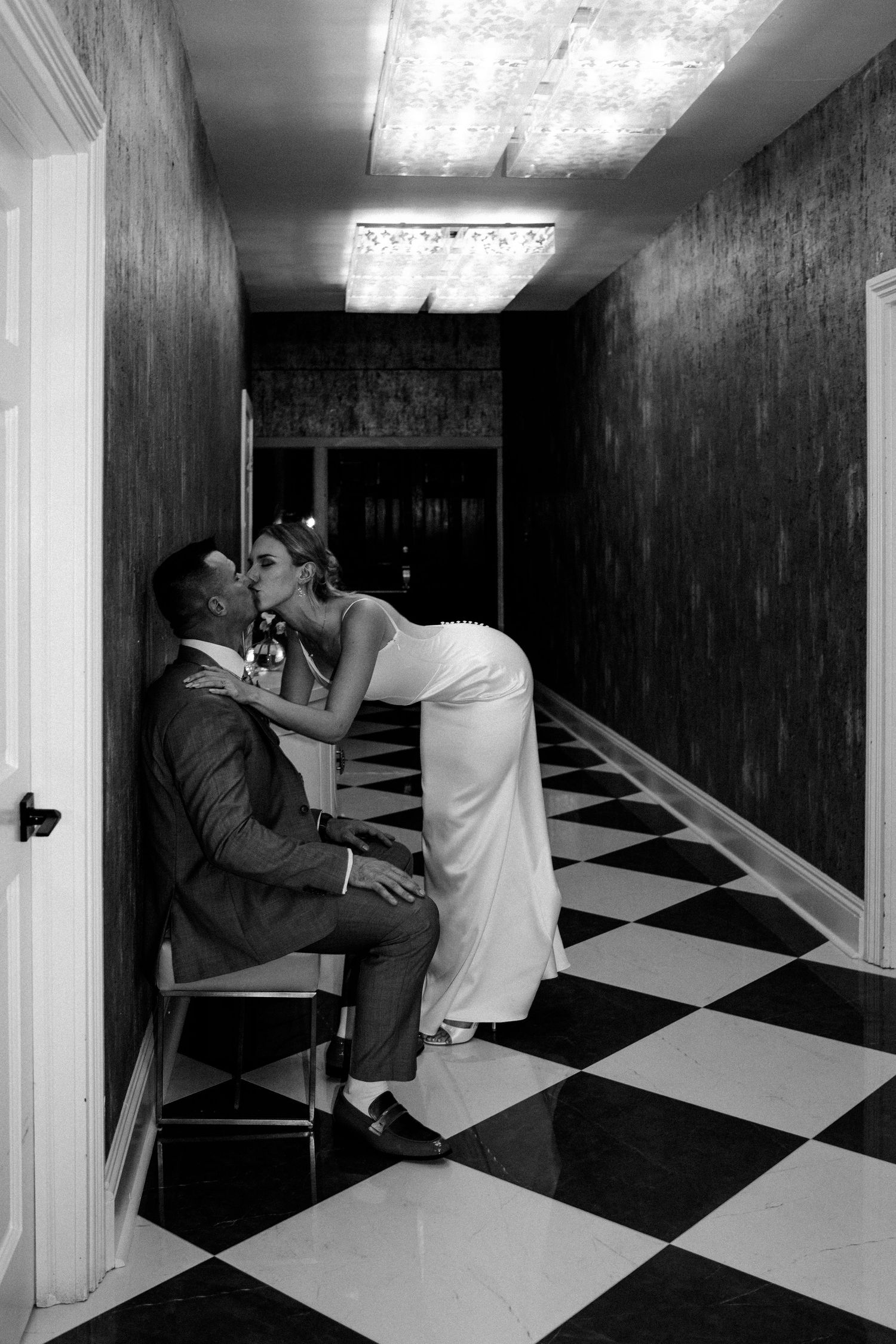 Black and white photo of bride and groom sharing a kiss in a checkered hallway.