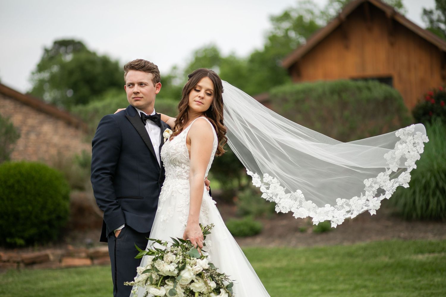 Bride and groom pose outdoors as her floral-trimmed veil billows in the wind, bride holding white bouquet.