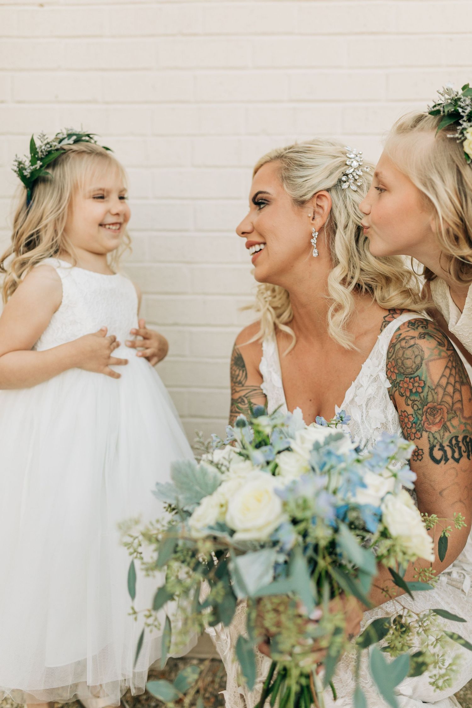 Two blonde brides in white lace gowns share a kiss near a flower girl in a white dress outdoors.