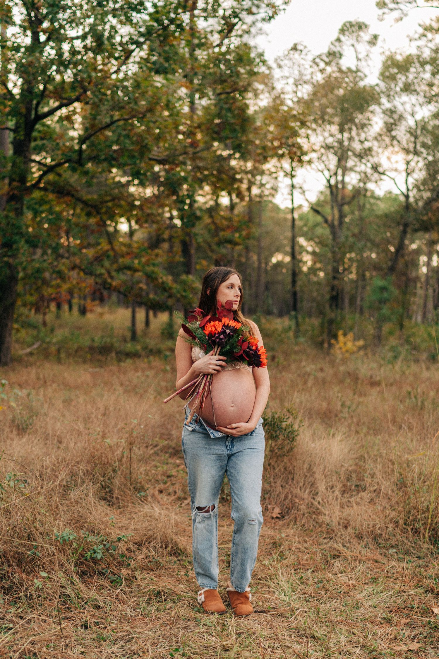 Pregnant woman in ripped jeans holding colorful autumn bouquet while standing in a sunlit forest clearing.