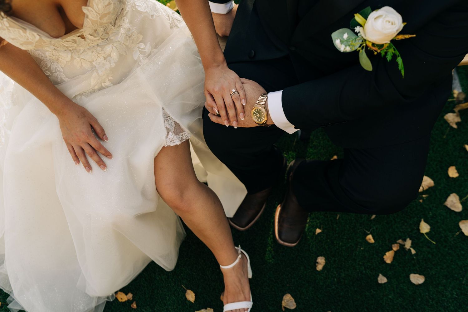 Bride and groom sitting together, holding hands, showcasing wedding rings, bridal gown, and boutonniere on green grass.