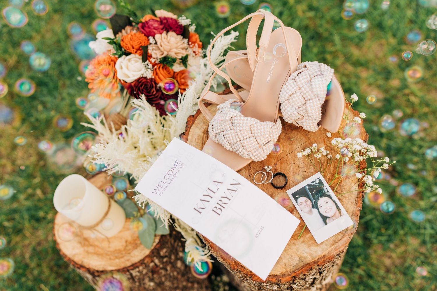Boho wedding flat lay featuring a vibrant bouquet, macrame heels, rings, and invitation on a wood stump.