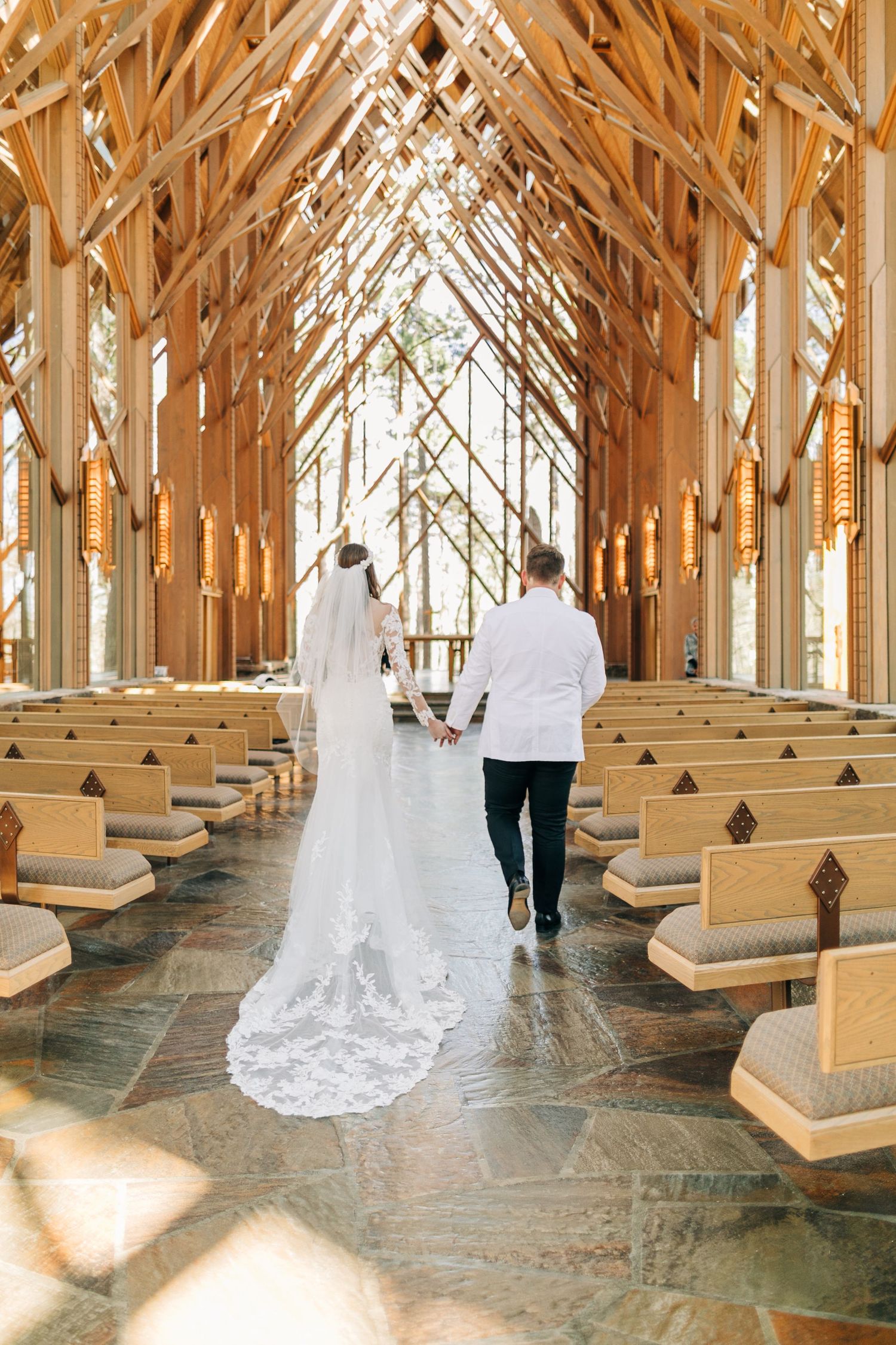 Bride and groom walking hand in hand down the aisle of a stunning wooden chapel with glass walls.