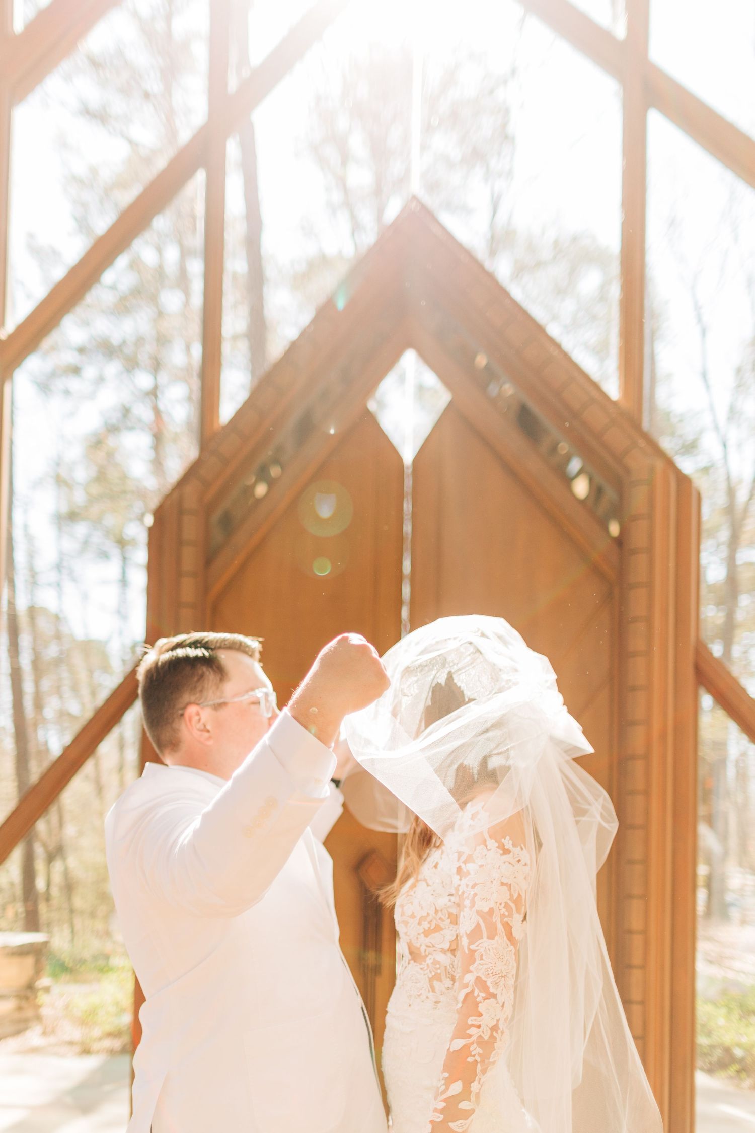 Groom lifts bride's veil in golden sunlight before stunning wooden chapel doors in forest setting.