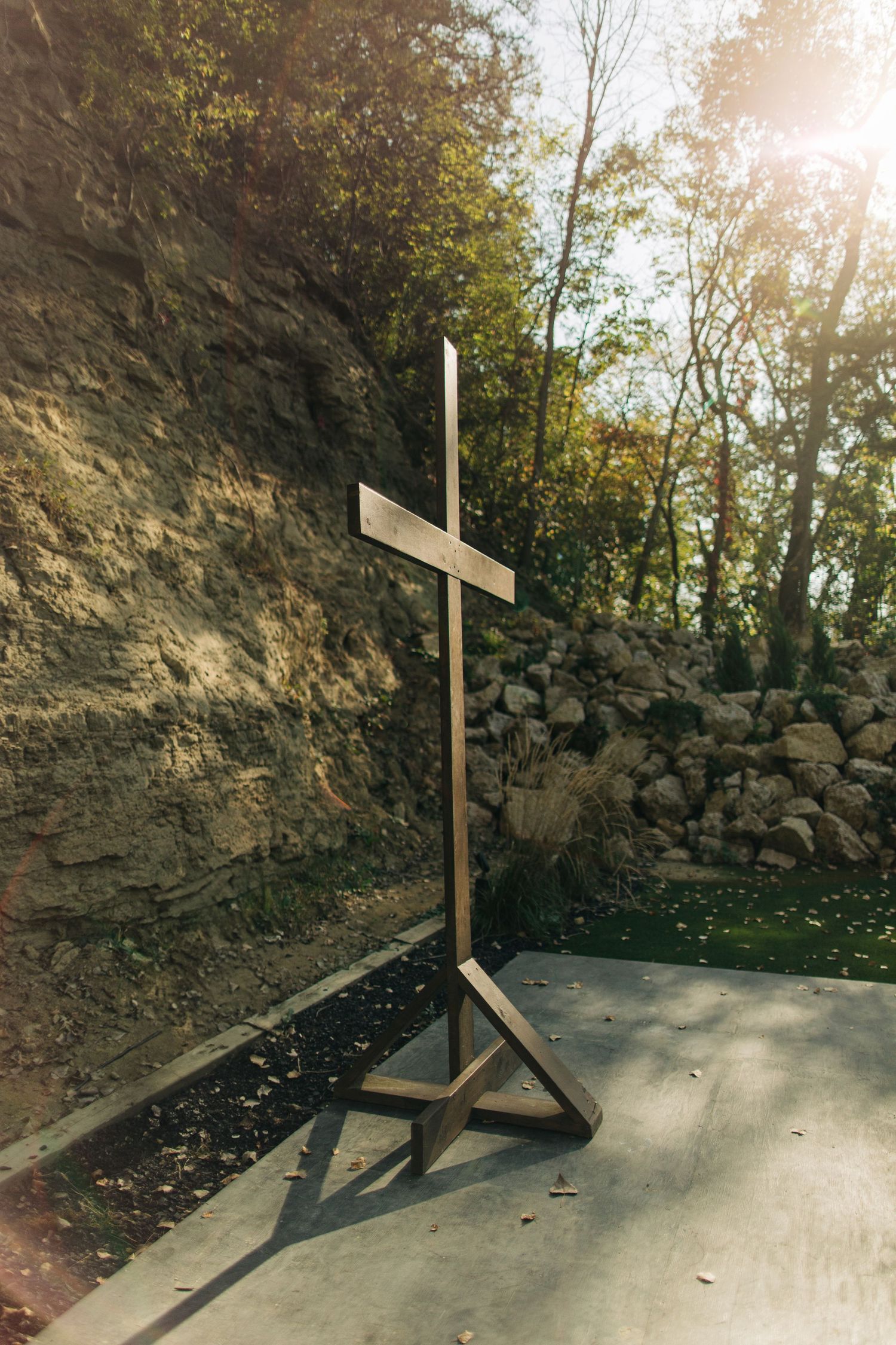 Rustic wooden cross standing outdoors against a sunlit rocky hillside with green trees.