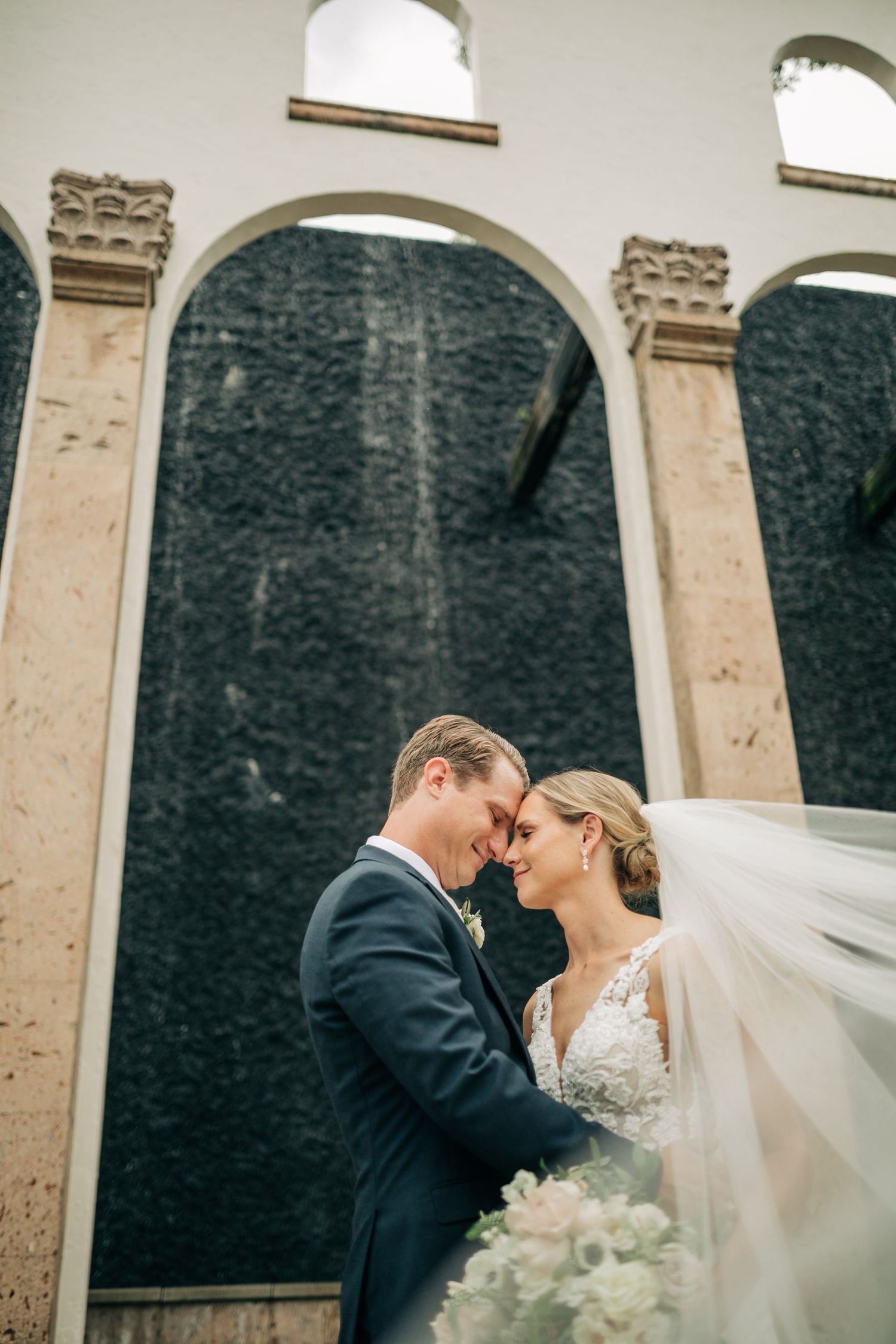 Romantic bride and groom touching foreheads, flowing veil billowing, standing before stone arched waterfall backdrop.