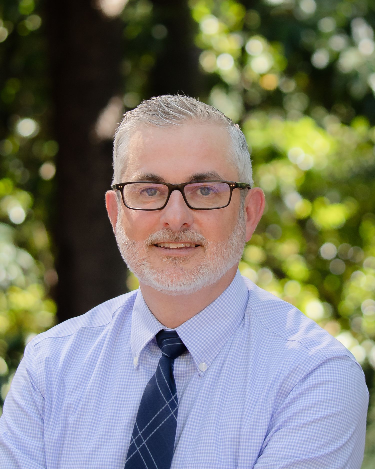 Professional headshot of someone in glasses wearing a light blue dress shirt and navy tie against a blurred outdoor background.