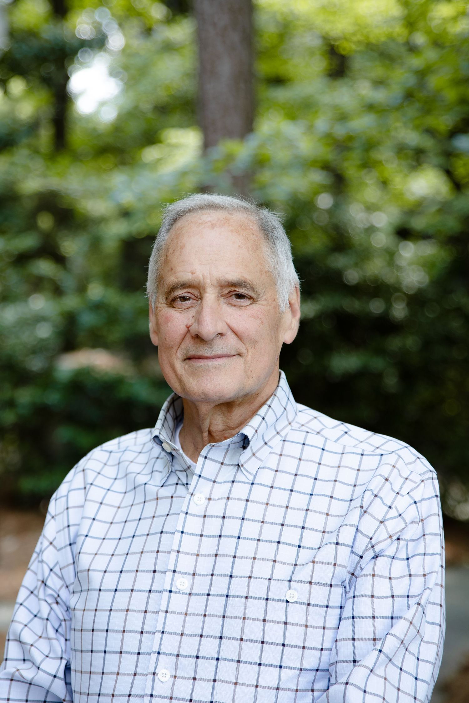 Smiling senior man with gray hair wearing a plaid shirt, standing outdoors surrounded by green trees.