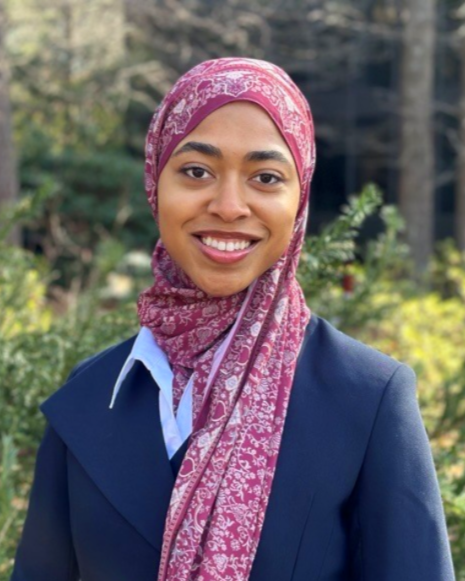 Young woman smiling in a red floral hijab and navy blazer outdoors with greenery in the background.