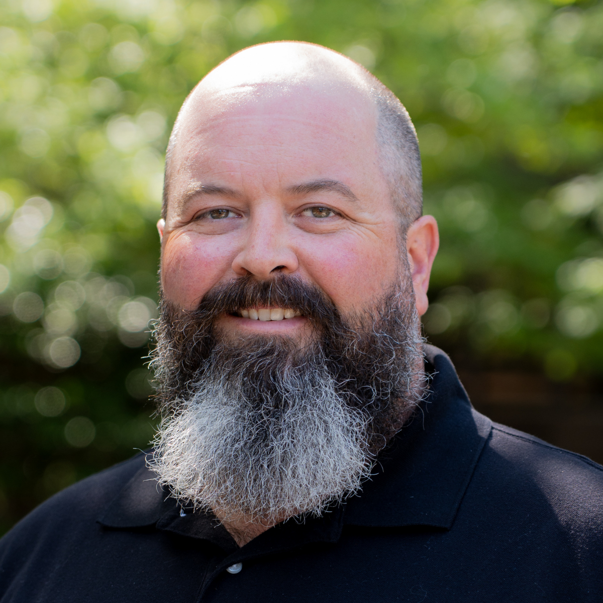 Smiling bald man with a full gray beard wearing a black shirt, photographed outdoors with green foliage background.