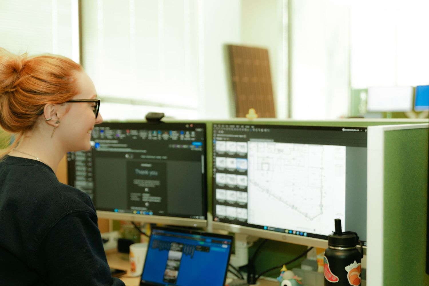 Red-haired woman with glasses working at dual monitor workstation displaying CAD architectural drawings in office.