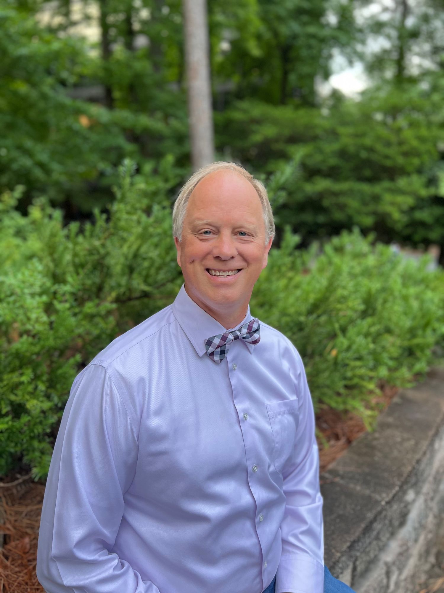 A person in a light blue dress shirt and bow tie sits outdoors with trees and greenery in the background.