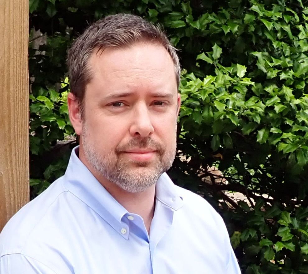 Professional headshot portrait of someone wearing a light blue button-down shirt against a green leafy background.