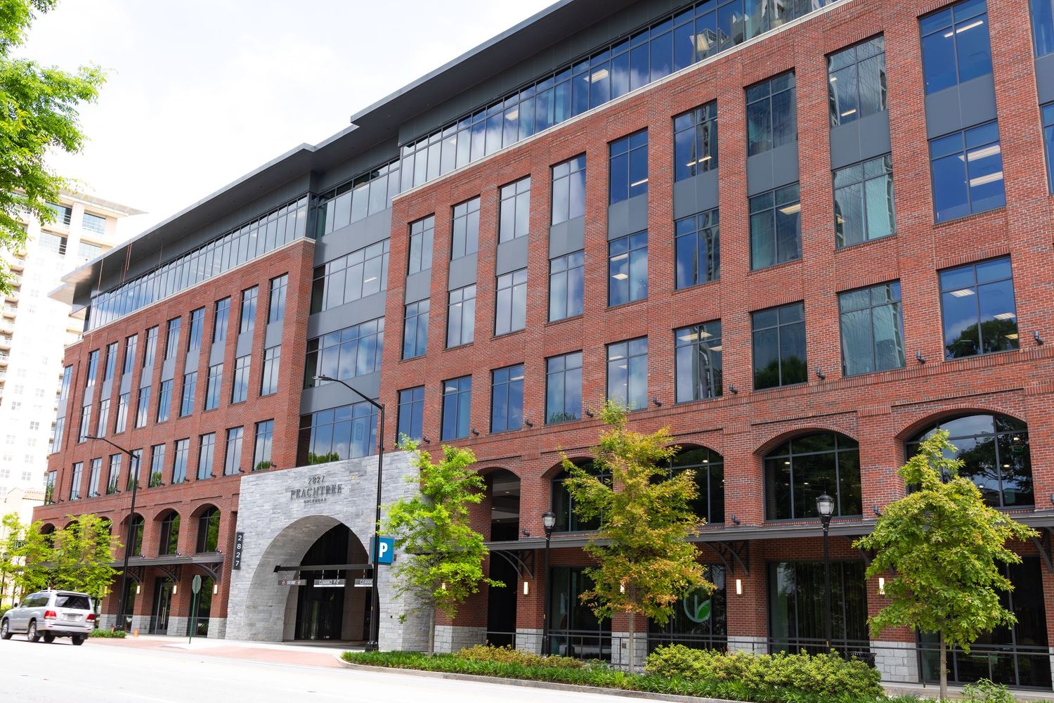 Modern red brick office building with glass windows and arched entryways along a tree-lined urban street.