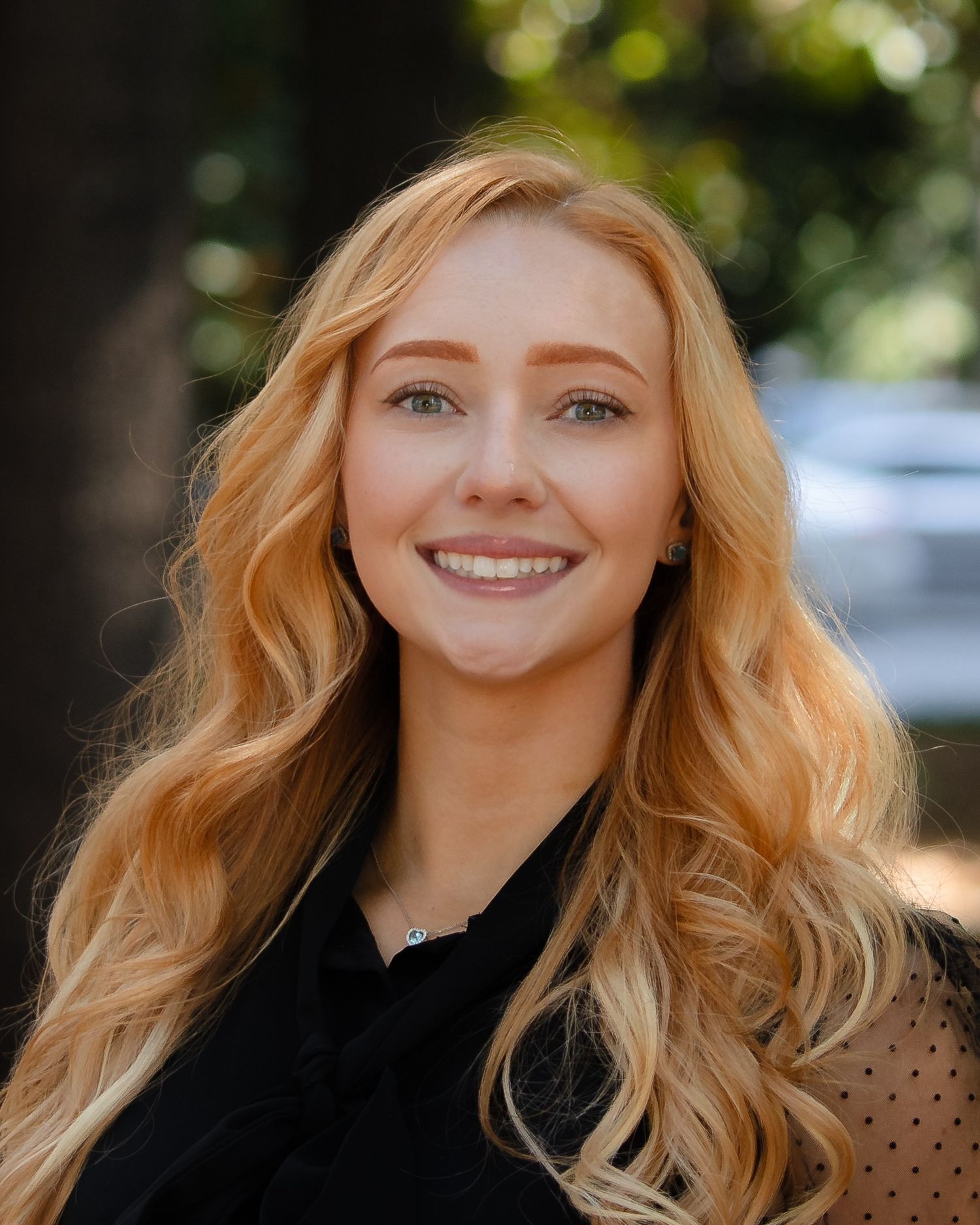 A young woman with wavy blonde hair smiles warmly at the camera in an outdoor setting with natural lighting.