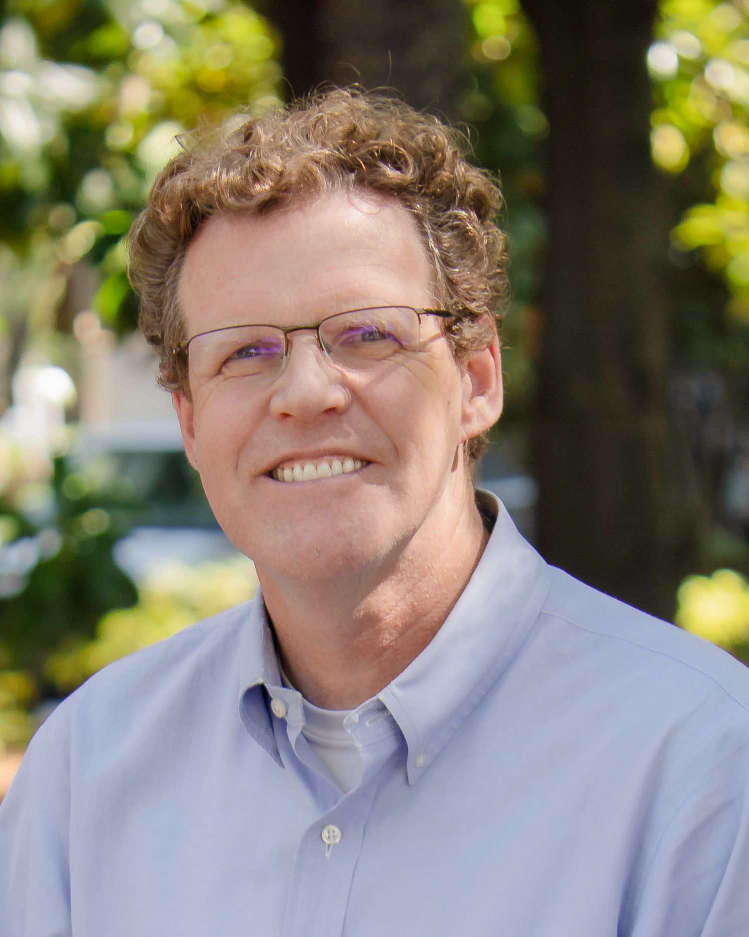 Professional headshot of someone wearing glasses and a light blue button-down shirt, photographed outdoors with greenery in background.