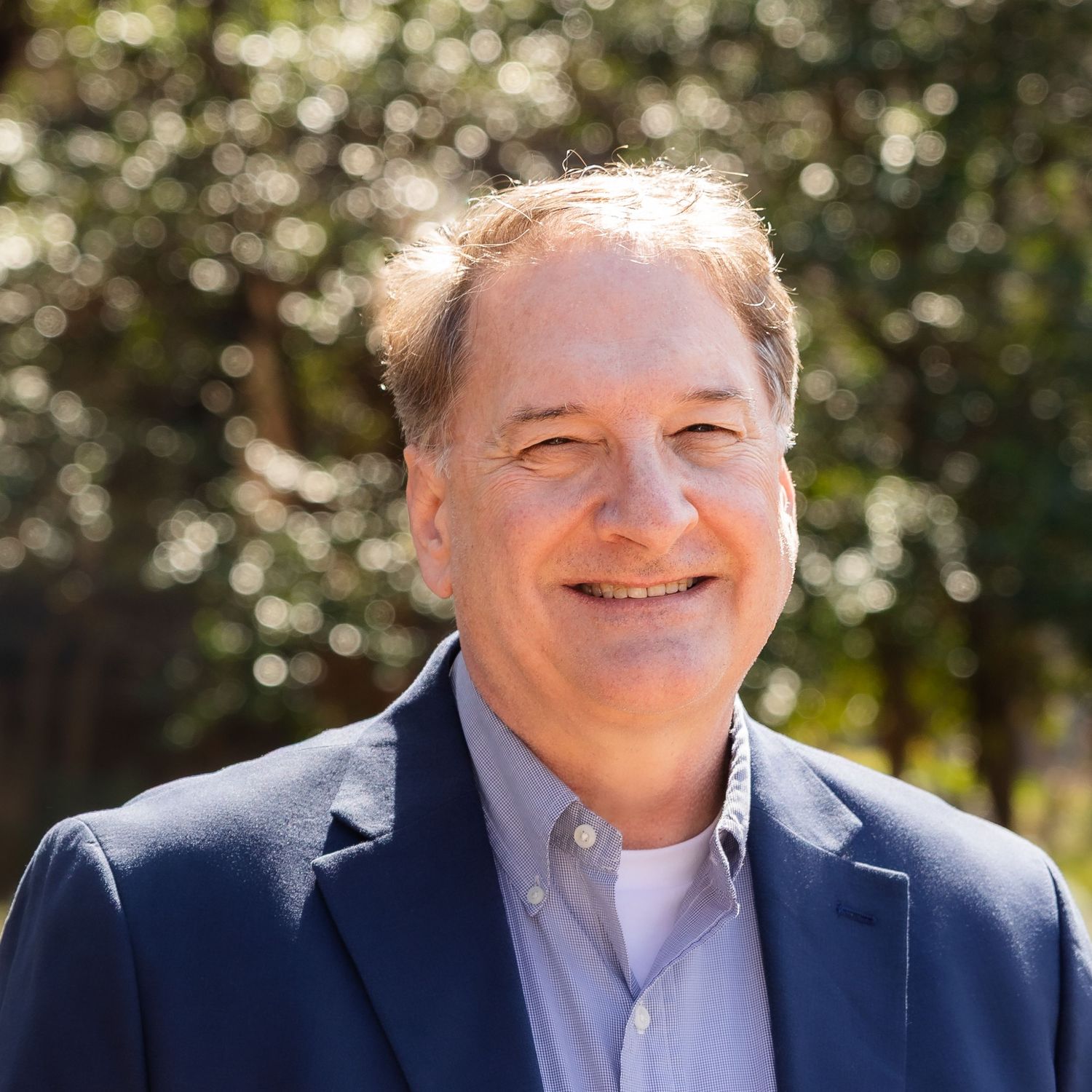 Person in navy suit jacket smiling warmly against natural outdoor background with sunlit foliage.
