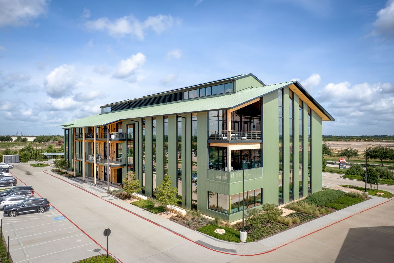 Modern green multi-story office building with wood accents, balconies, and landscaped surroundings under a cloudy sky.